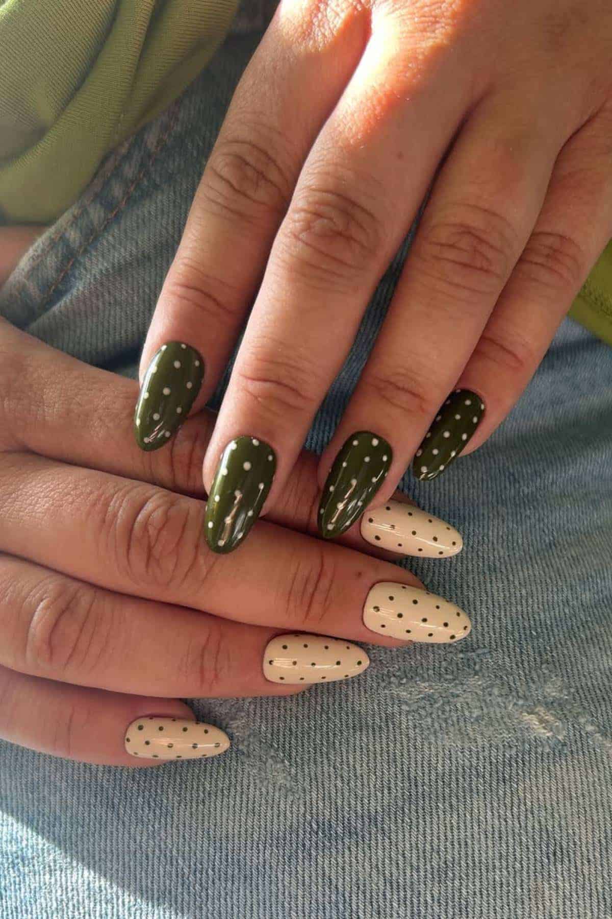 Two hands with almond-shaped nails; one hand has dark green nails with white polka dots, the other has white nails with black polka dots, resting on blue denim.