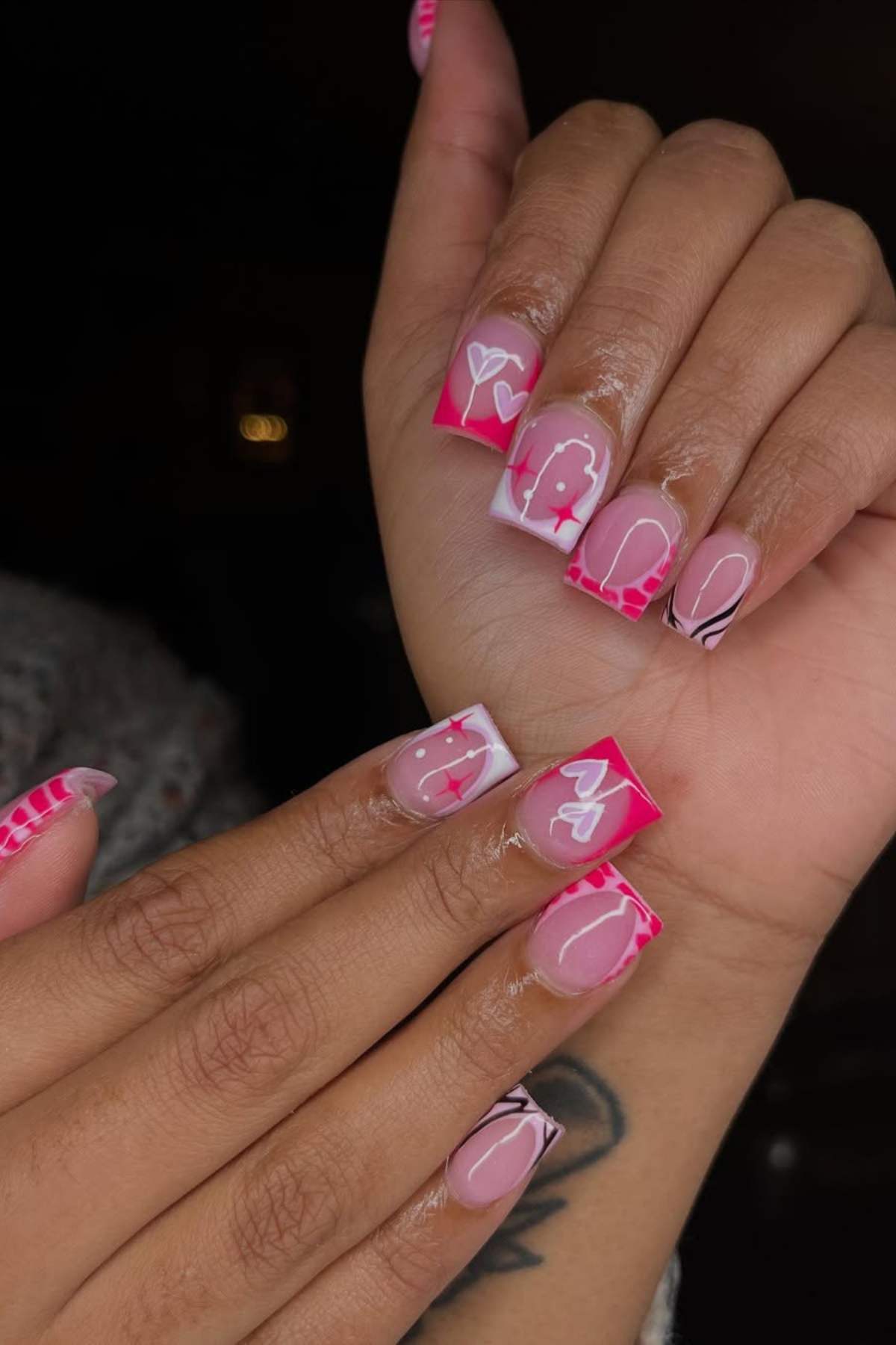A person with medium-length square acrylic nails featuring pink, red, and white designs, including hearts, swirls, dots, and animal print accents.