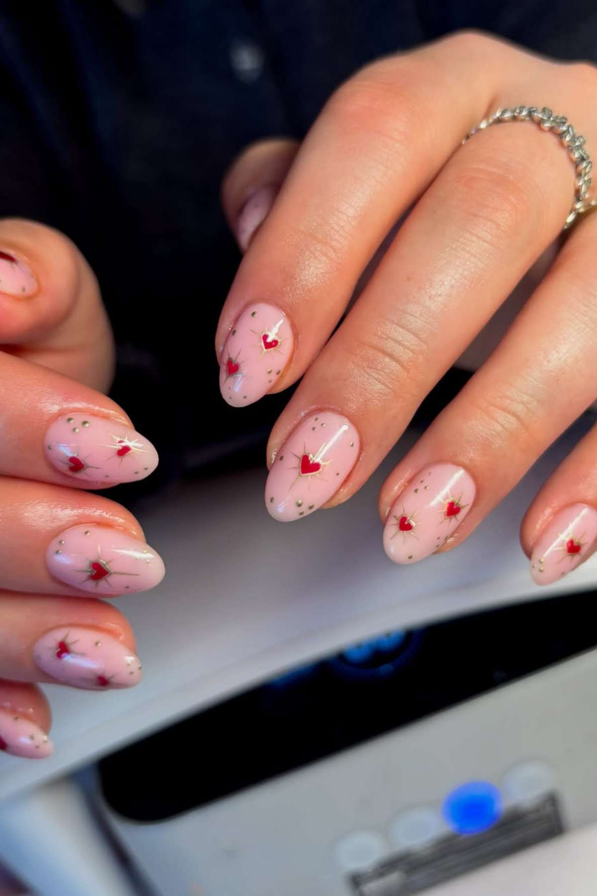 Close-up of hands with almond-shaped nails painted light pink, decorated with small red hearts and silver specks.