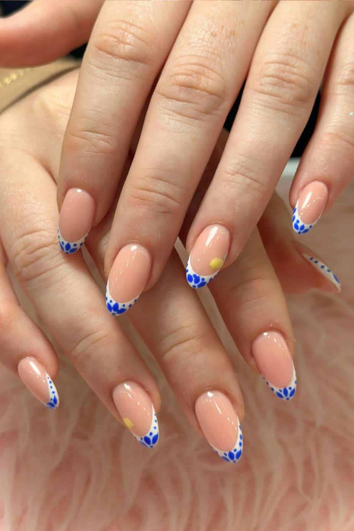 Hands with almond-shaped nails featuring nude polish, blue heart-shaped tips, and small yellow accent dots on some nails.