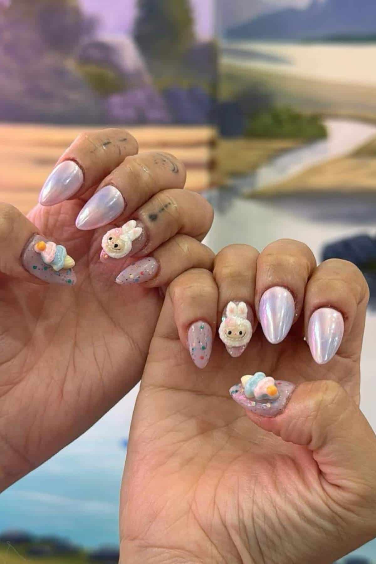 Hands with long, almond-shaped nails featuring iridescent polish and 3D bunny and ice cream cone designs, held in front of a blurred outdoor-themed background.