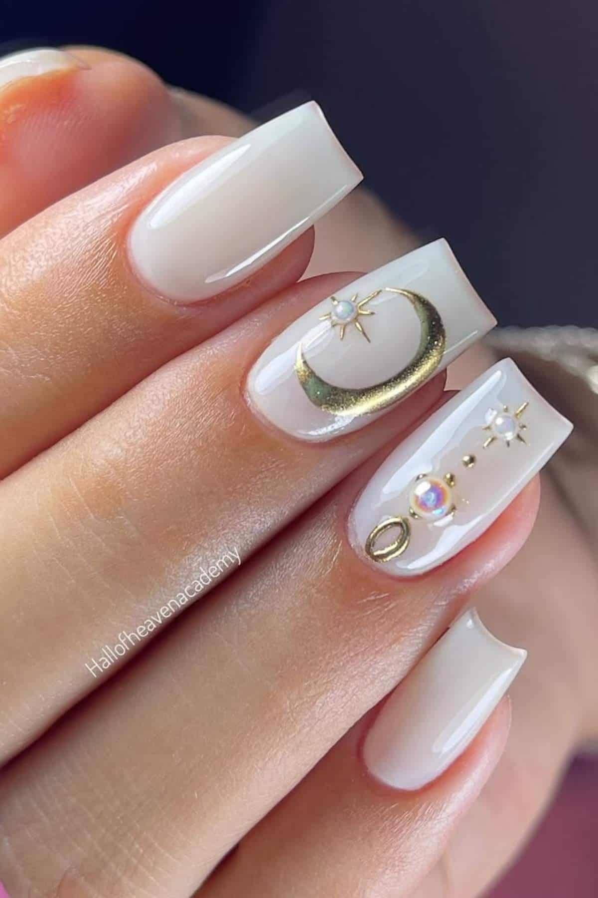 A hand with square-shaped, glossy nude nails featuring gold celestial designs, including a crescent moon, stars, and small gems on two accent nails.
