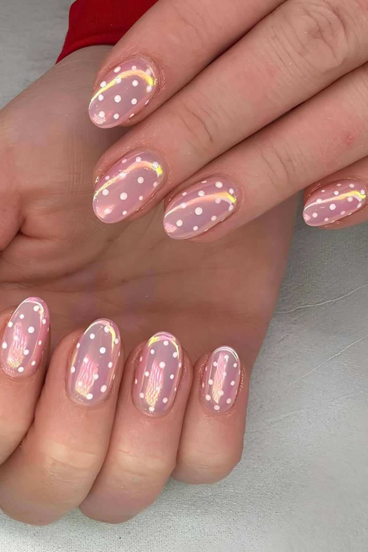 A hand with oval-shaped nails painted light pink, decorated with white polka dots and a thin, curved gold line on each nail.