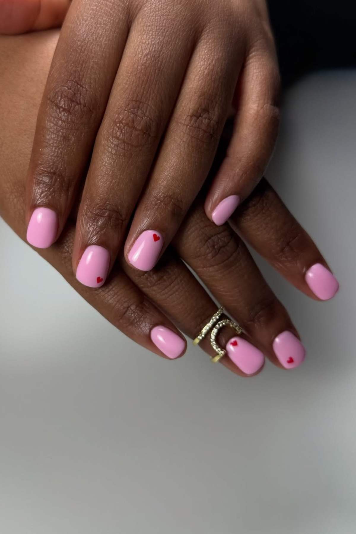 Two hands with short, square-shaped nails painted light pink and decorated with small red heart accents; one hand wears a gold ring with a rhinestone letter "C.