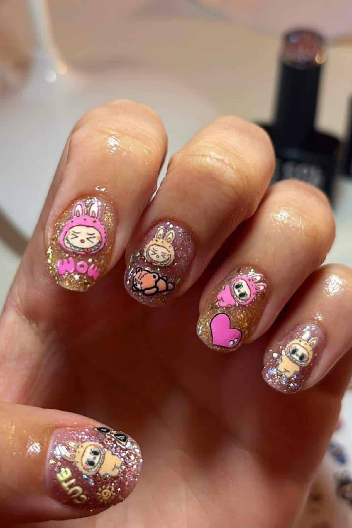 A hand with short nails decorated with glittery polish and cartoon character stickers, including hearts, animals, and text designs.