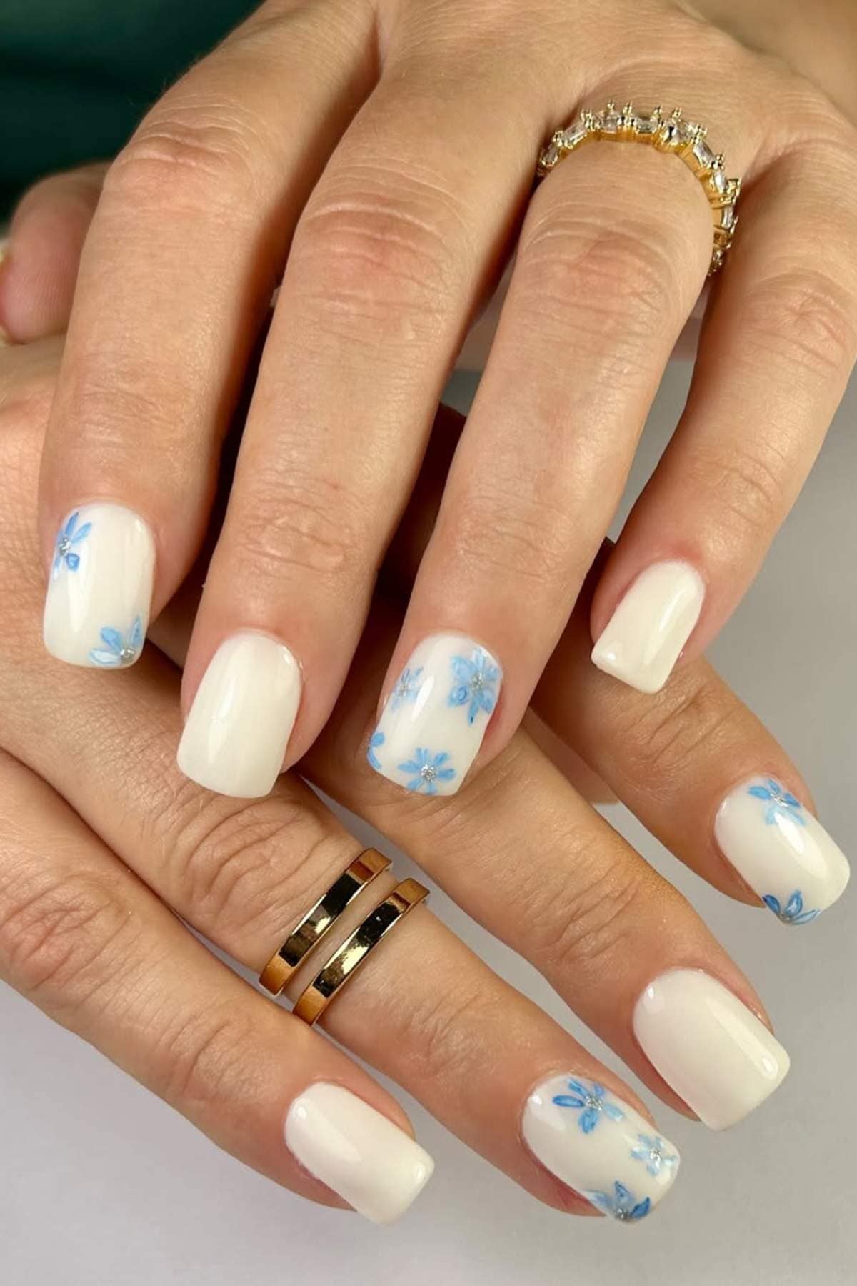 Hands with square-shaped white nails featuring small blue flower designs on some nails, and gold rings on two fingers.