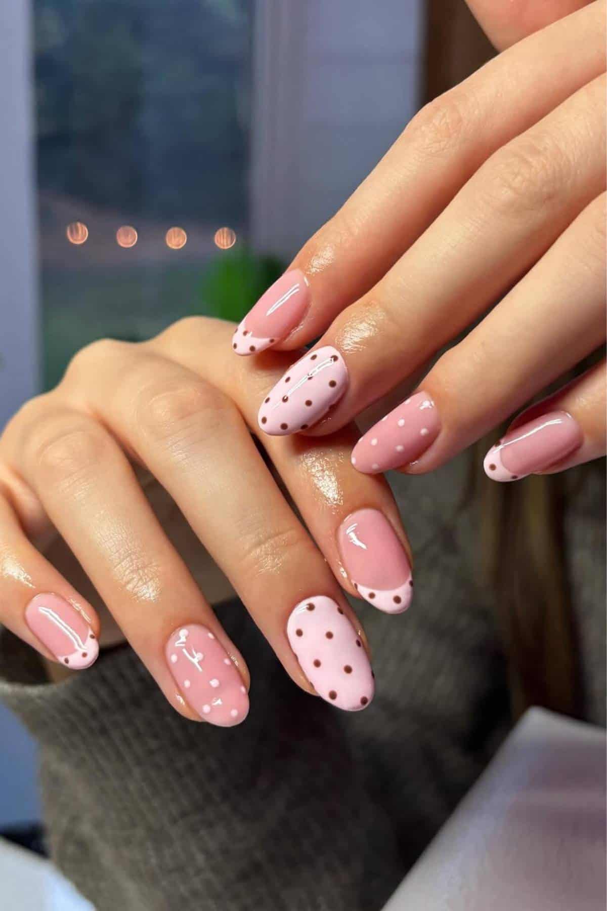 A close-up of hands with pink manicured nails featuring white and pink polka dots, stripes, and French tip designs.