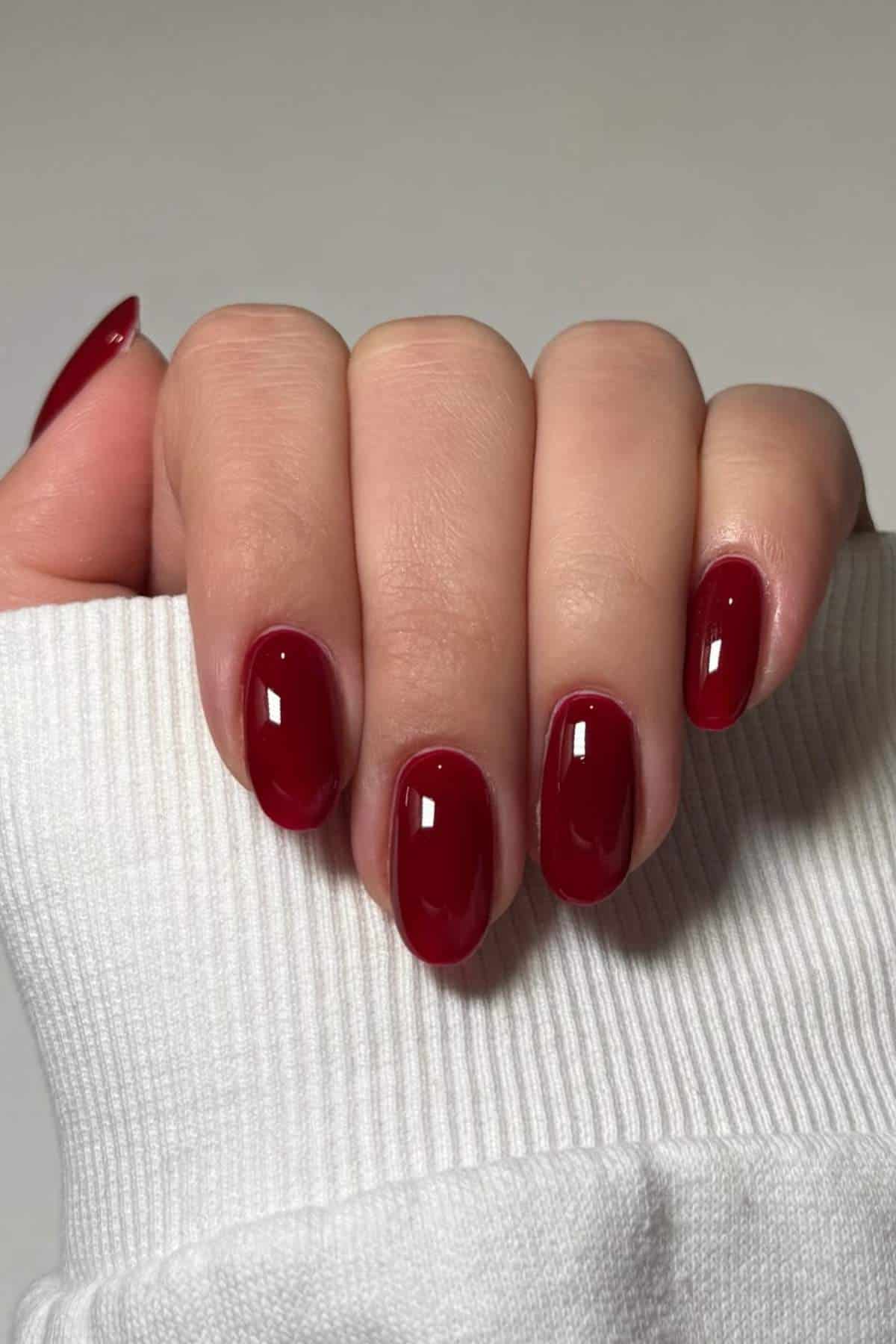 A hand with short, oval-shaped nails painted with glossy dark red nail polish, shown against a neutral background.