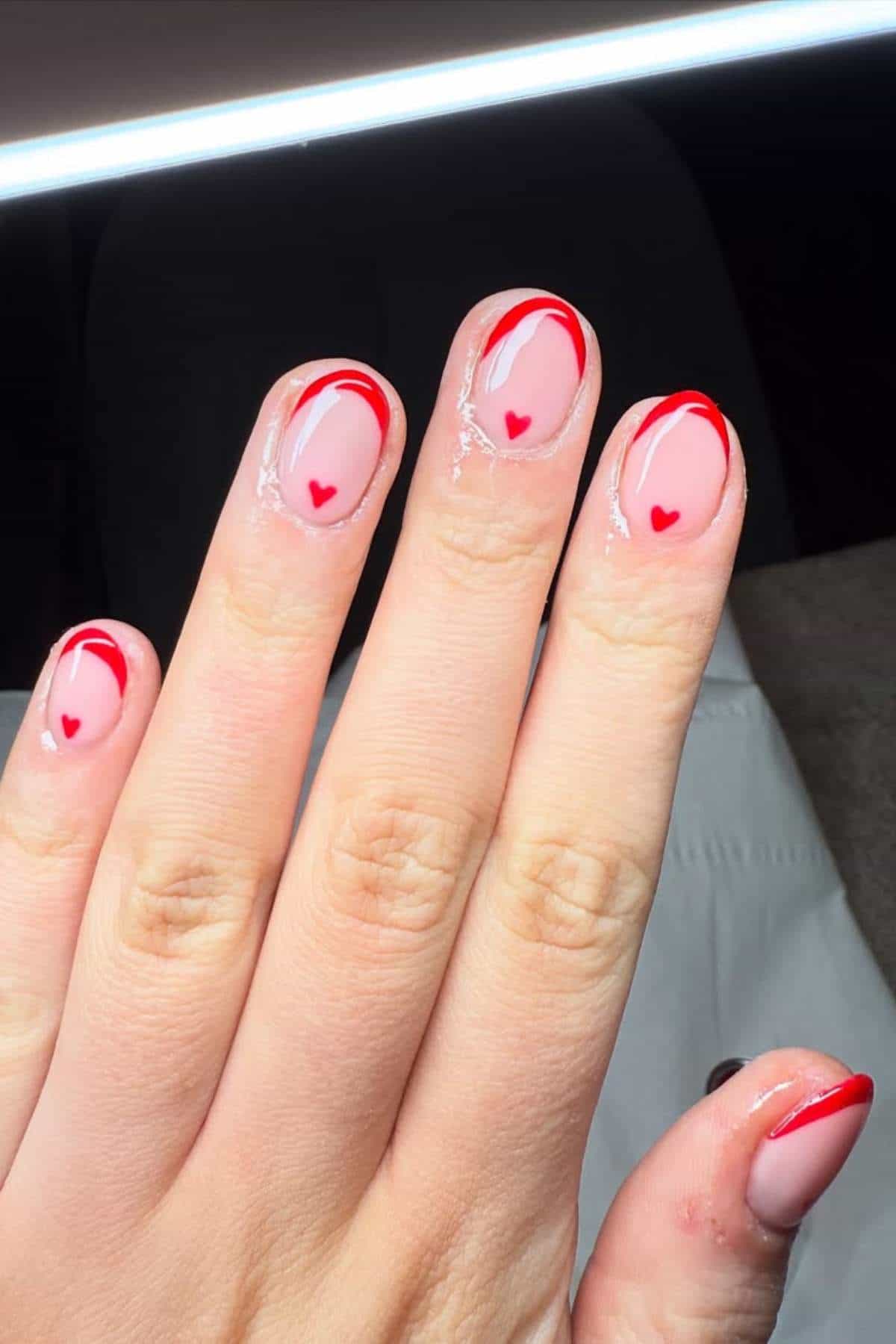 A hand with pink nails featuring red hearts near the cuticles and red curved tips, set against a neutral background.