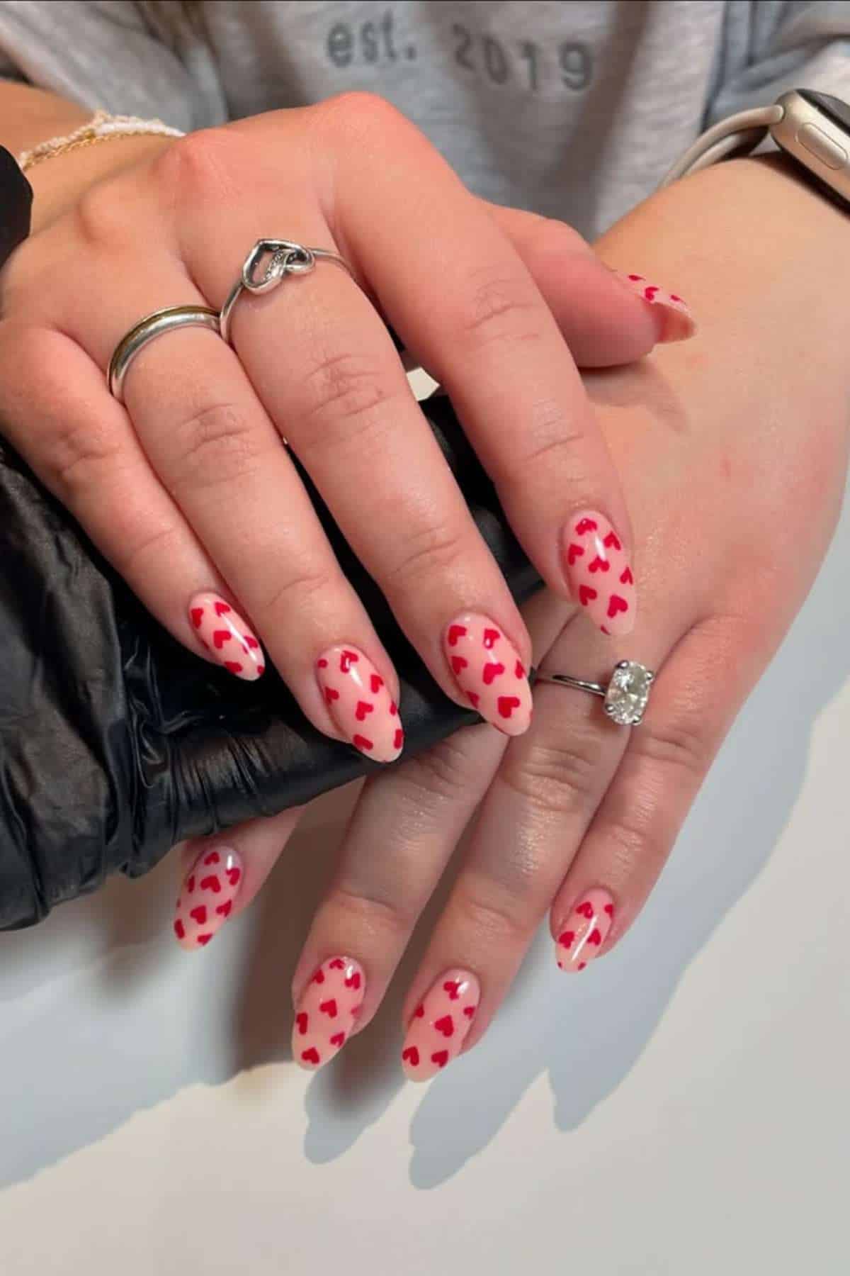 Two hands with almond-shaped nails painted light pink and decorated with small red hearts. The person is wearing rings and a smartwatch.