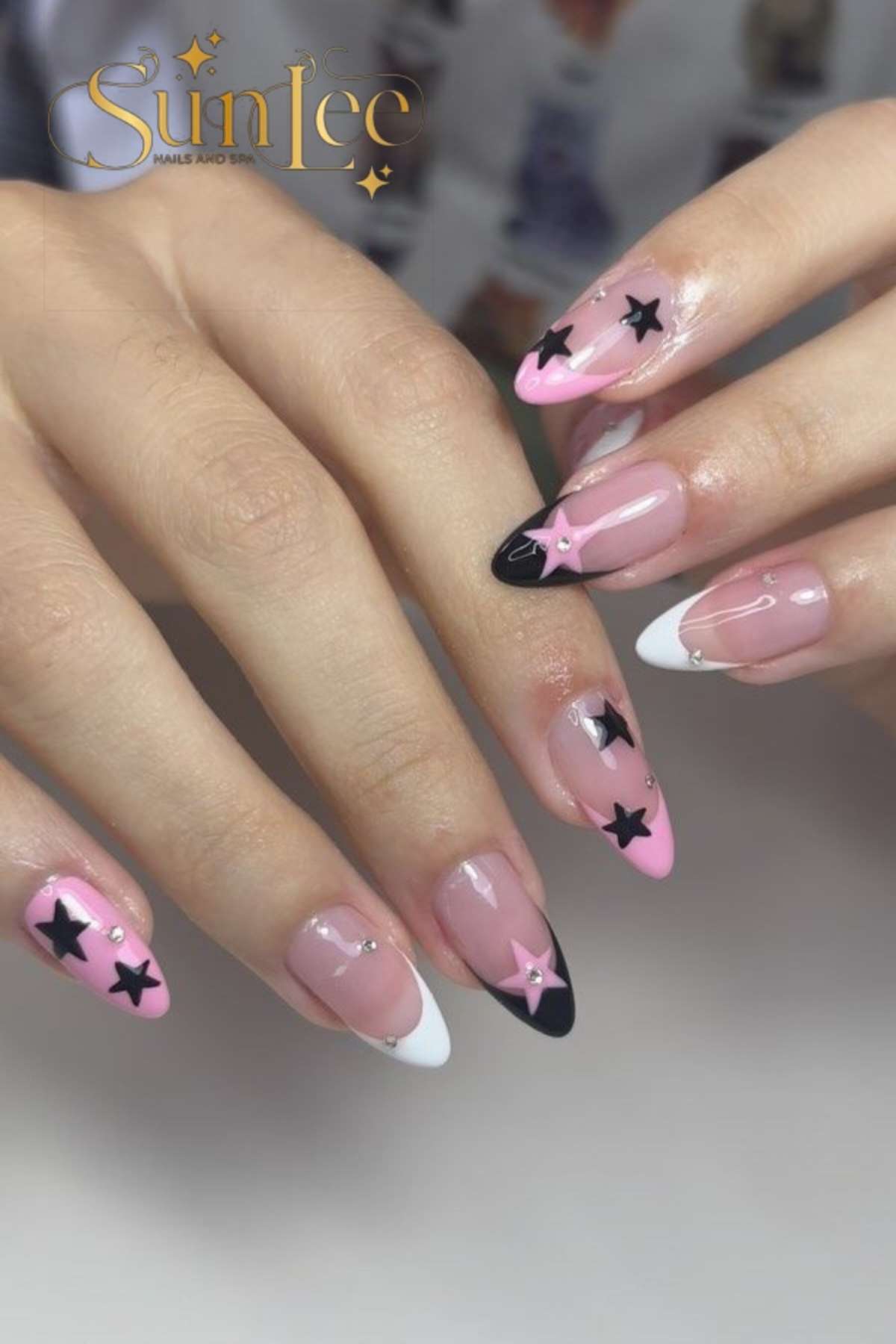 A hand with almond-shaped nails featuring pink, black, and white tips, decorated with black and pink star designs on a transparent base.