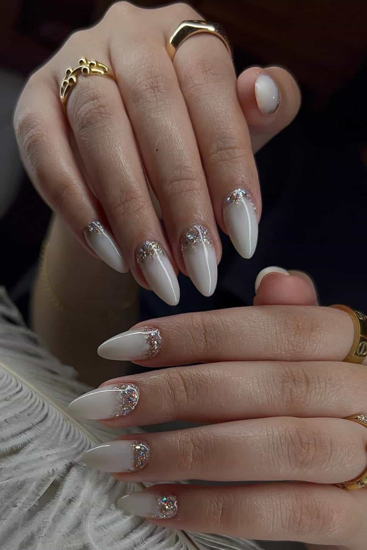 Hands with long, almond-shaped white nails featuring silver glitter accents near the cuticles, wearing gold rings.