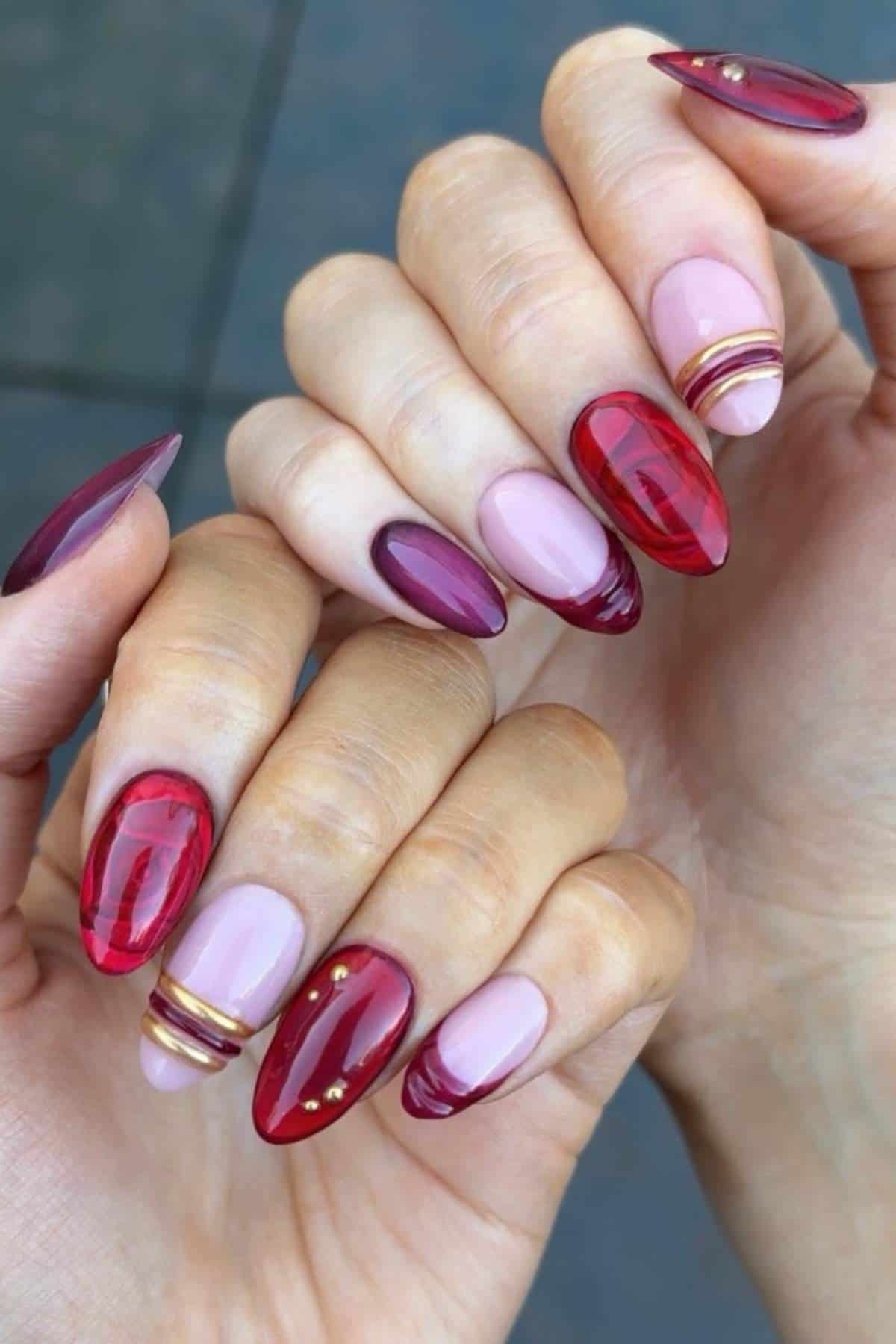 Hands with almond-shaped nails featuring a mix of translucent red, mauve, and nude polish, accented with gold rings and small gold studs.