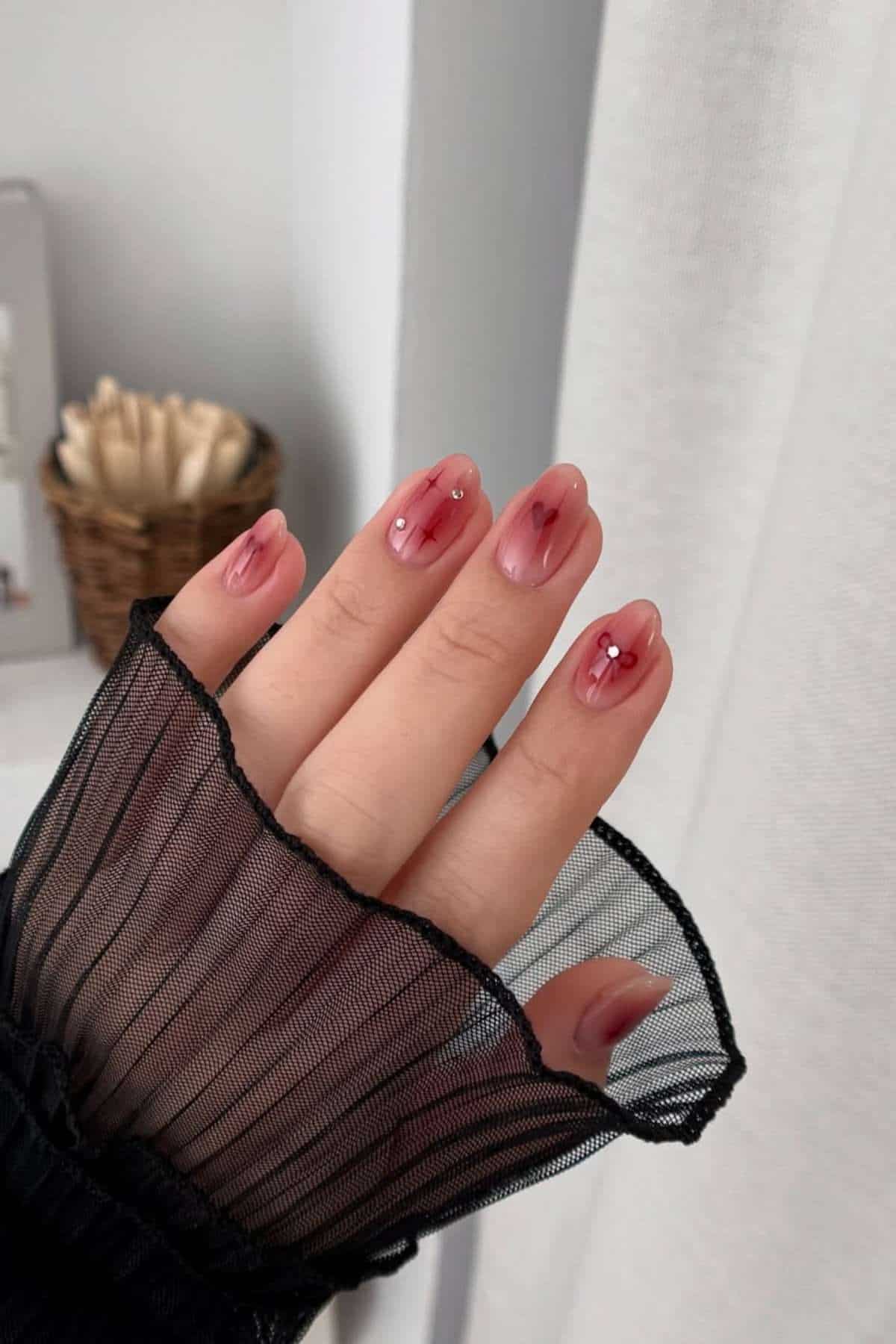 A hand with almond-shaped nails, featuring a pink ombre design, delicate white lines, and small rhinestone accents. The sleeve is black and sheer.