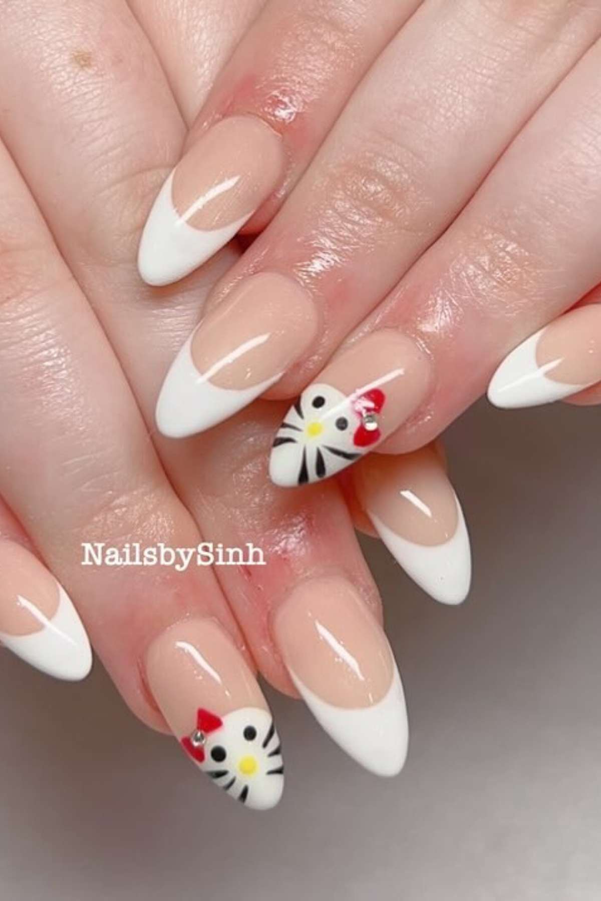 A hand with almond-shaped French tip nails; two nails feature a cartoon cat face with a red bow and whiskers.