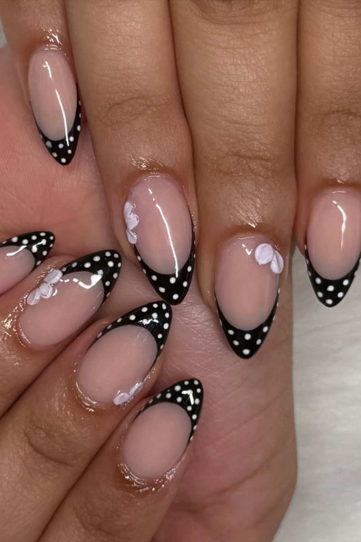 A hand with almond-shaped nails featuring black French tips with white polka dots and small 3D white floral accents on a nude base.