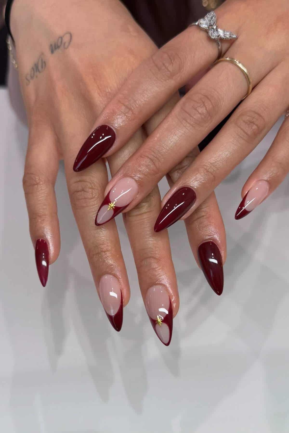 Hands with long, almond-shaped nails featuring a deep red polish, nude and red French tips, and small gold embellishments on two nails.