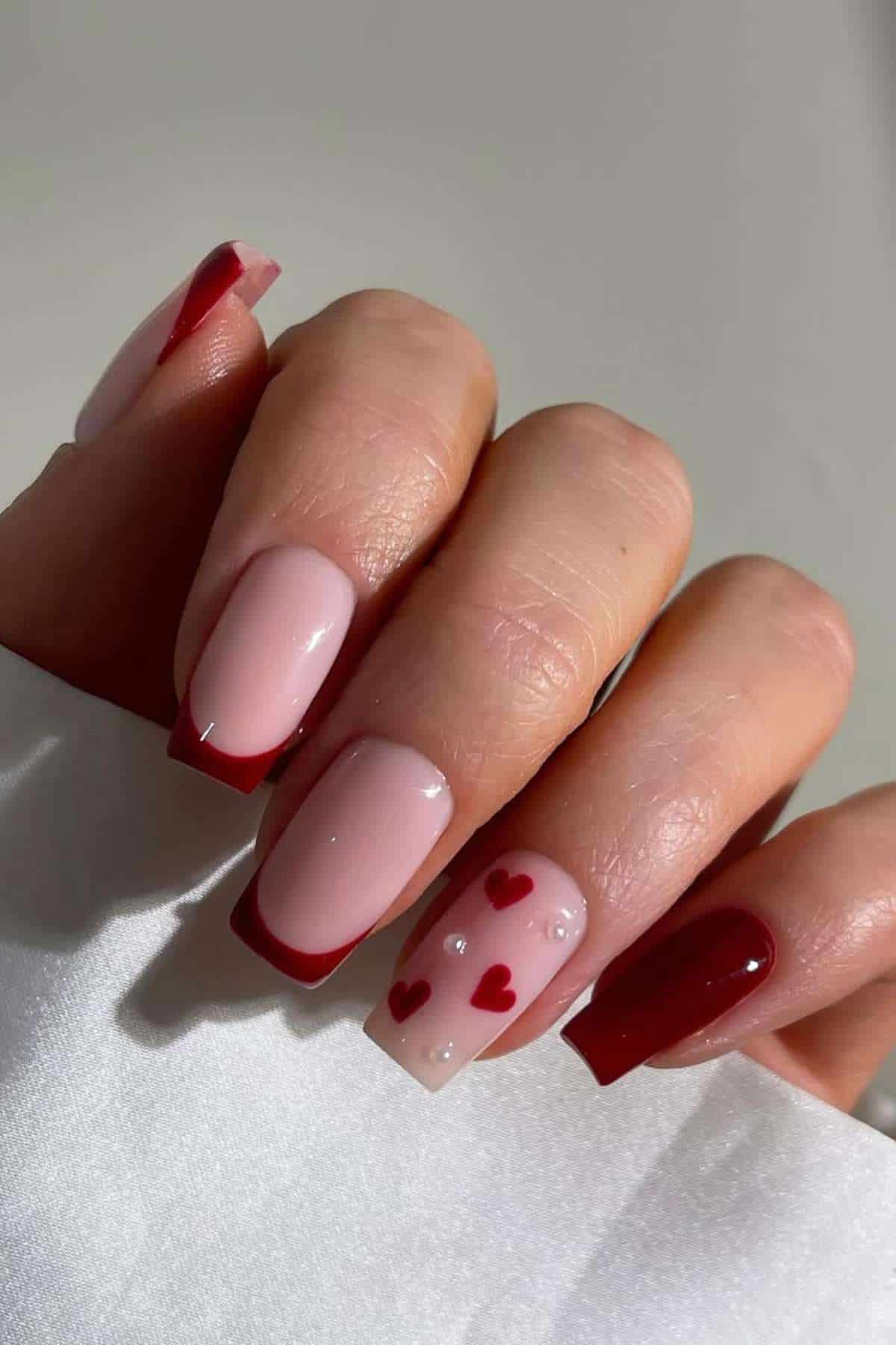 A hand with square acrylic nails; some nails are painted red, others are nude with red French tips, and one features red hearts and small white dots on a nude base.