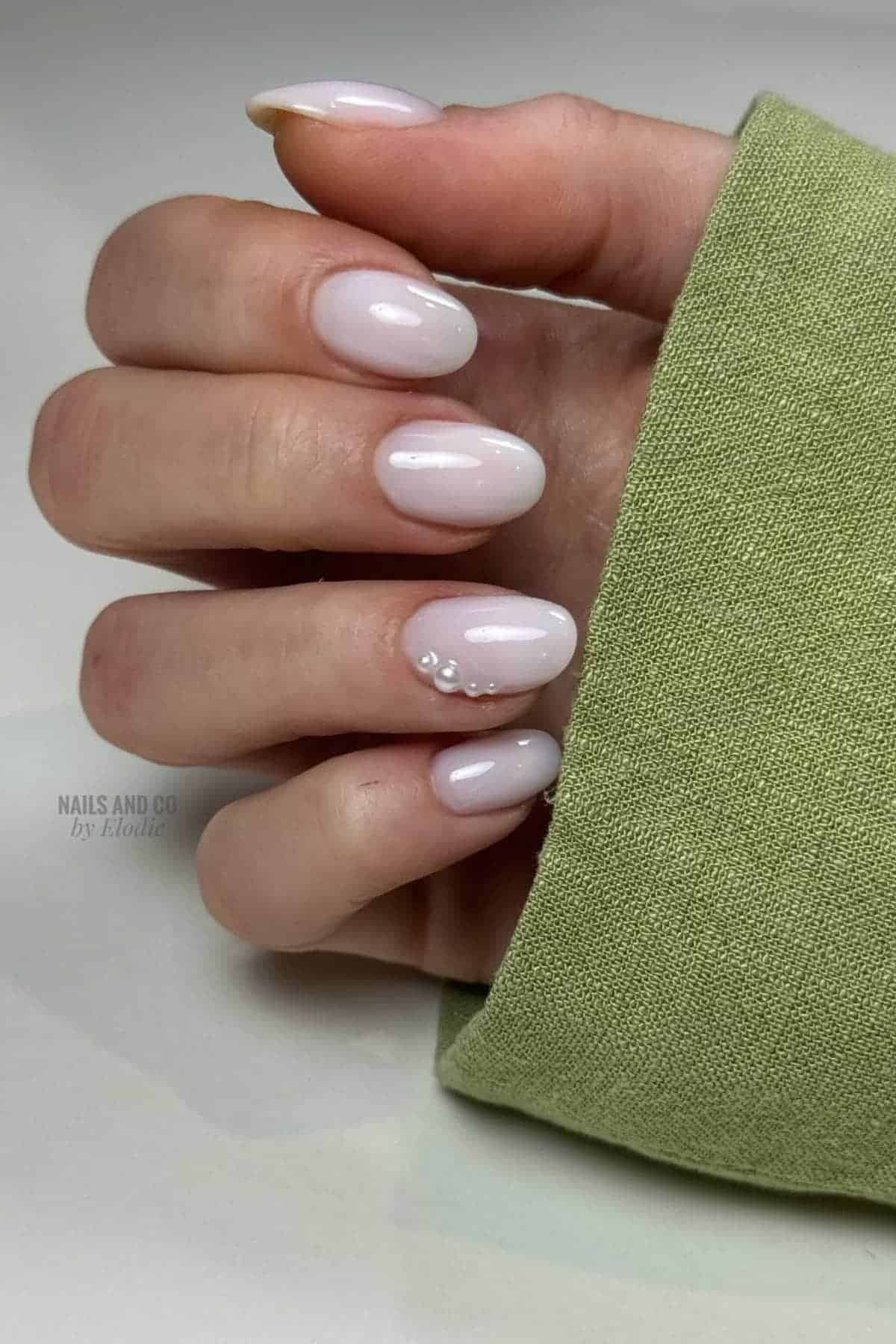 A hand with short, oval-shaped, pale pink nails; one nail decorated with small white pearls. The person is wearing a light green sleeve.