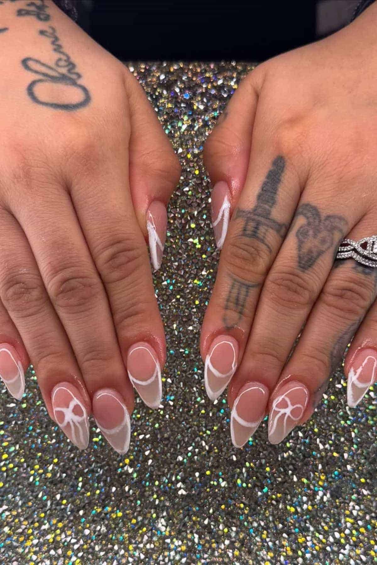 Hands with long, almond-shaped nails featuring white abstract line art designs, resting on a sparkly, glittery surface; visible tattoos on fingers and hands.