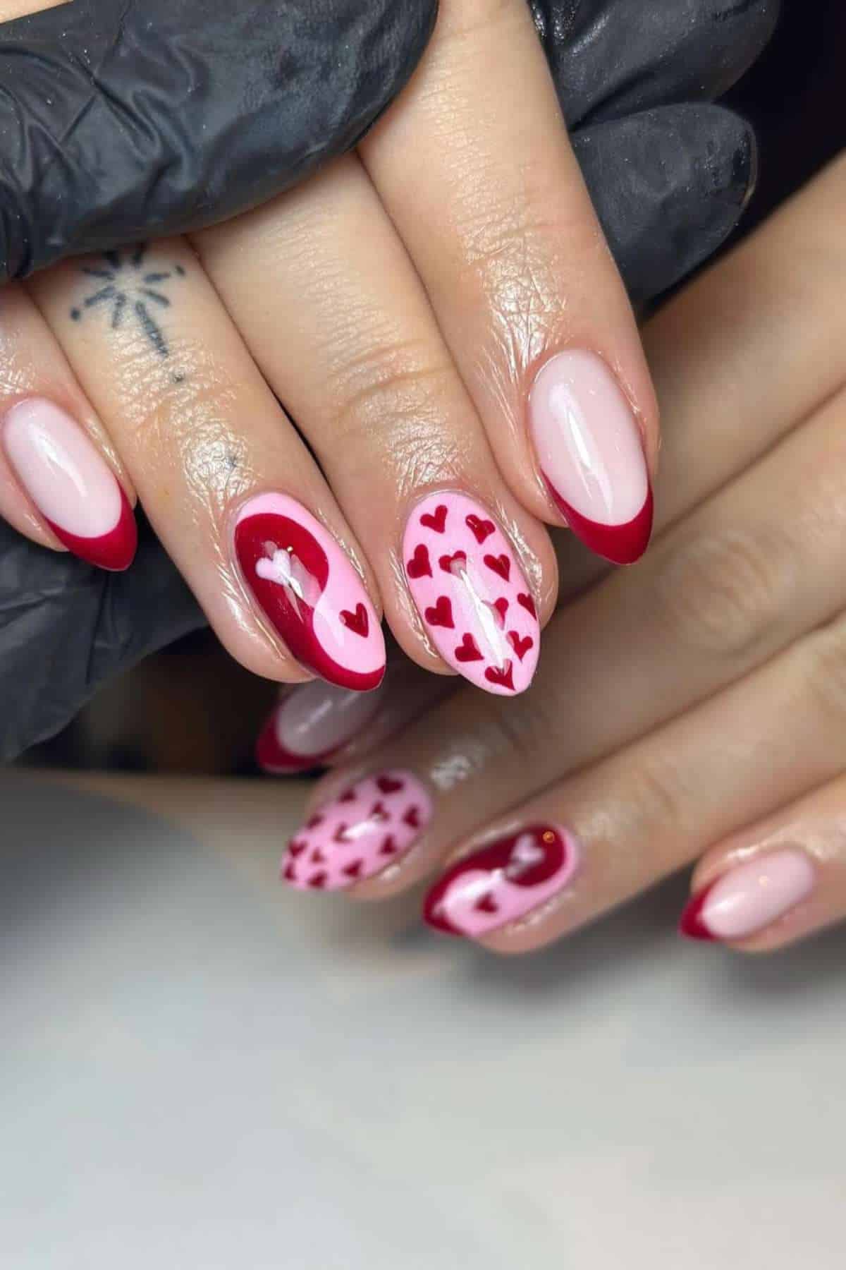 A hand with almond-shaped nails featuring red French tips and heart-themed nail art in red and pink tones. A gloved hand holds the fingers.