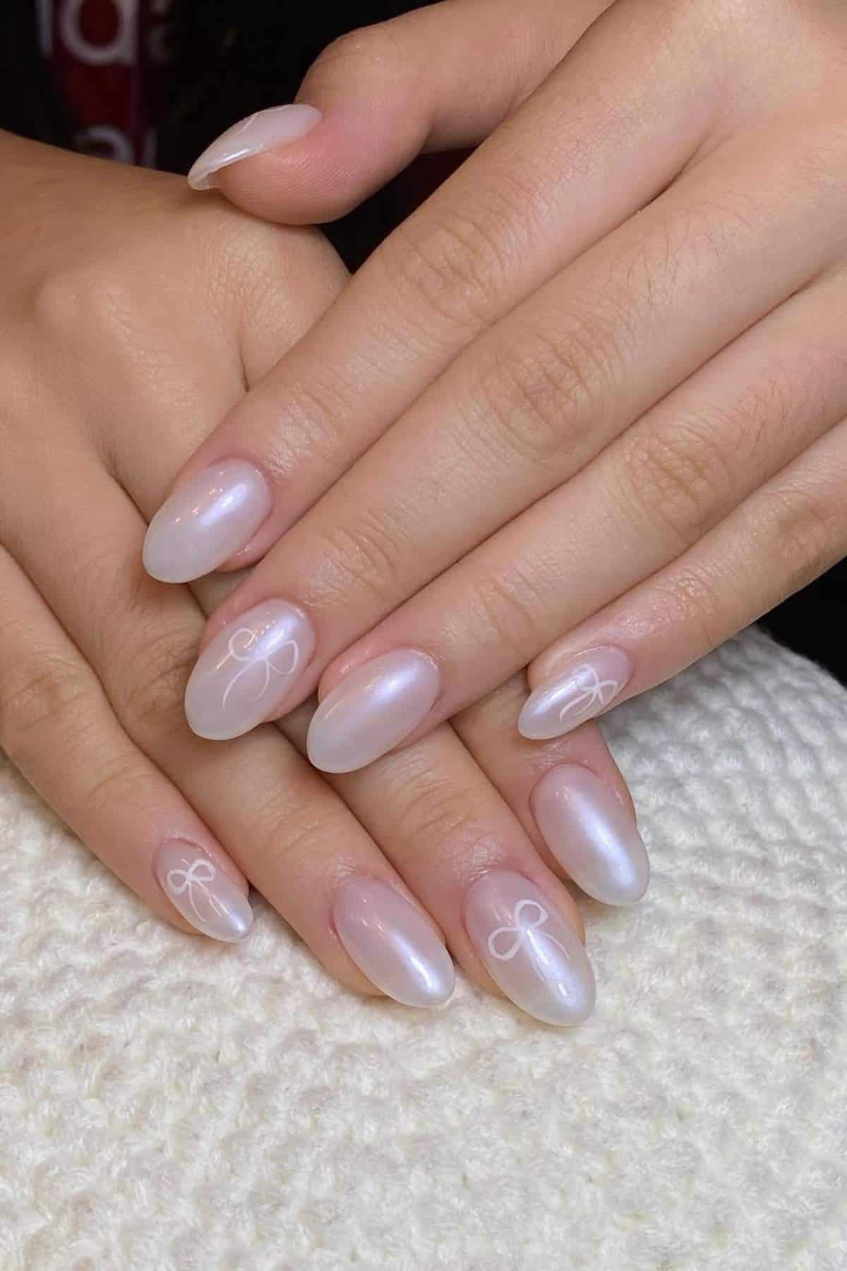 Hands with oval-shaped, pearly white manicured nails featuring small, delicate white bow designs on a few nails.