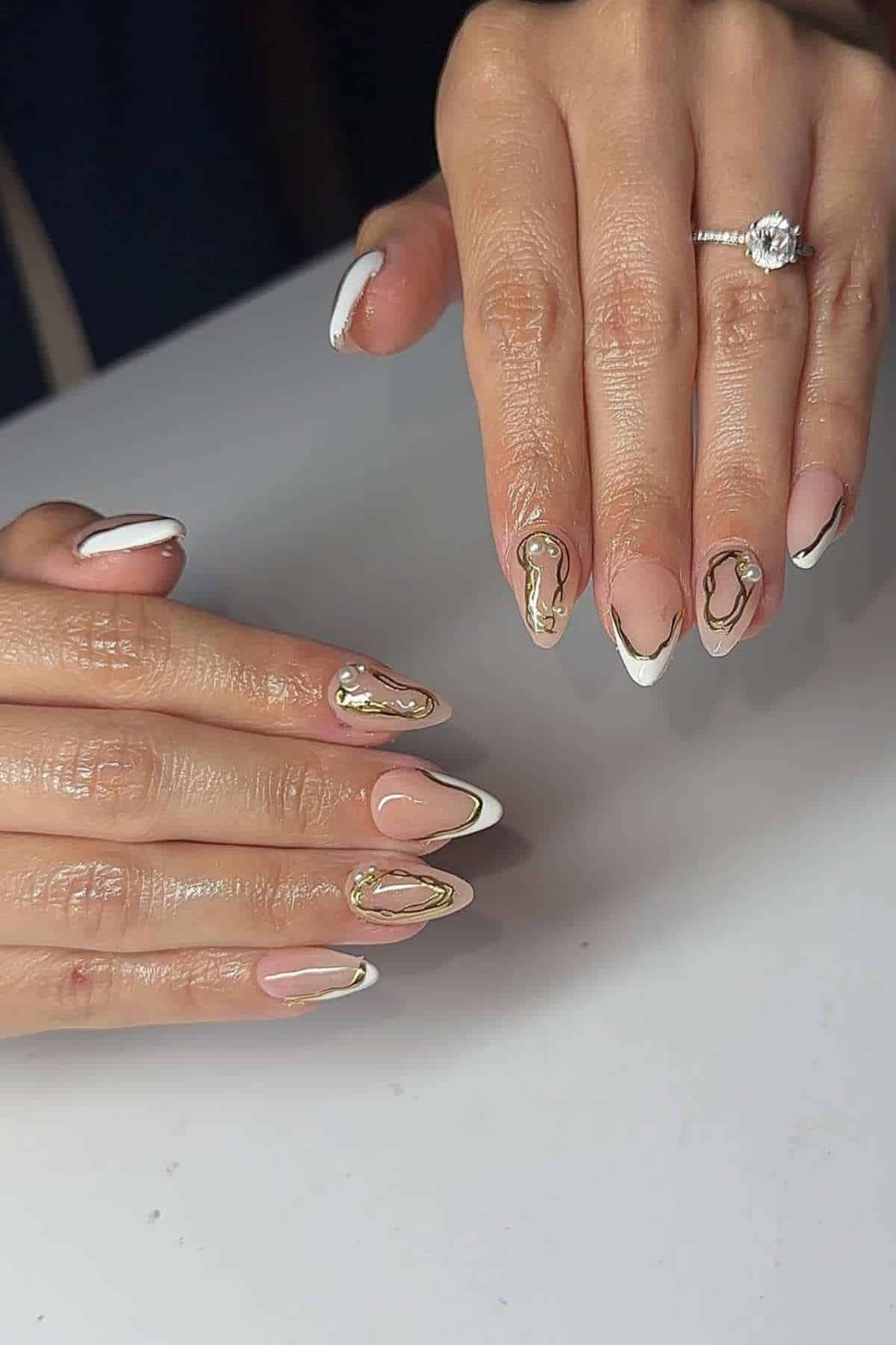 Hands with almond-shaped nails featuring nude polish, white and gold abstract designs, and a diamond ring on the ring finger of the right hand.