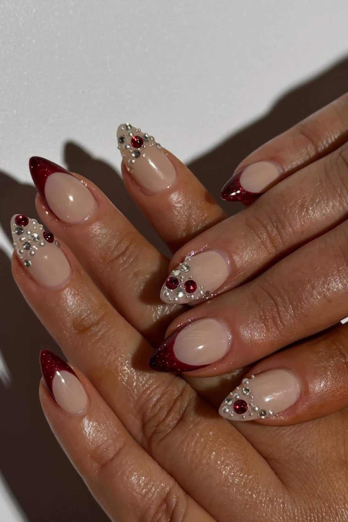 Hands with almond-shaped nails featuring nude polish, dark red tips, and decorative pearls and red gem accents on some nails.