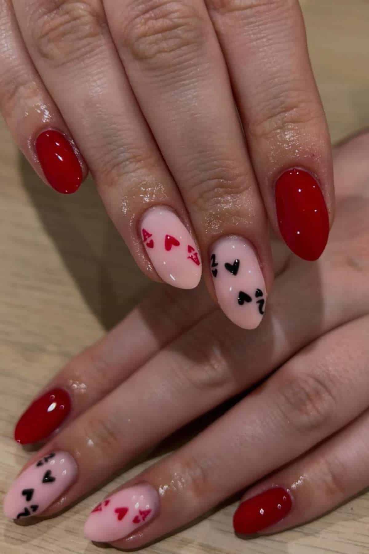 Hands with red and nude nails; nude nails feature small red and black playing card suit symbols, including hearts, spades, clubs, and diamonds.