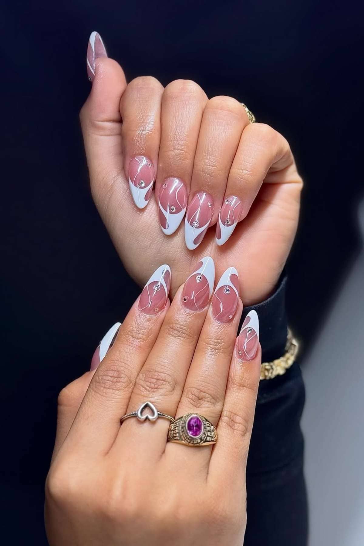 Close-up of hands showing almond-shaped nails with a clear base, white abstract tips, and small silver rhinestones; wearer has a gold bracelet and two rings.