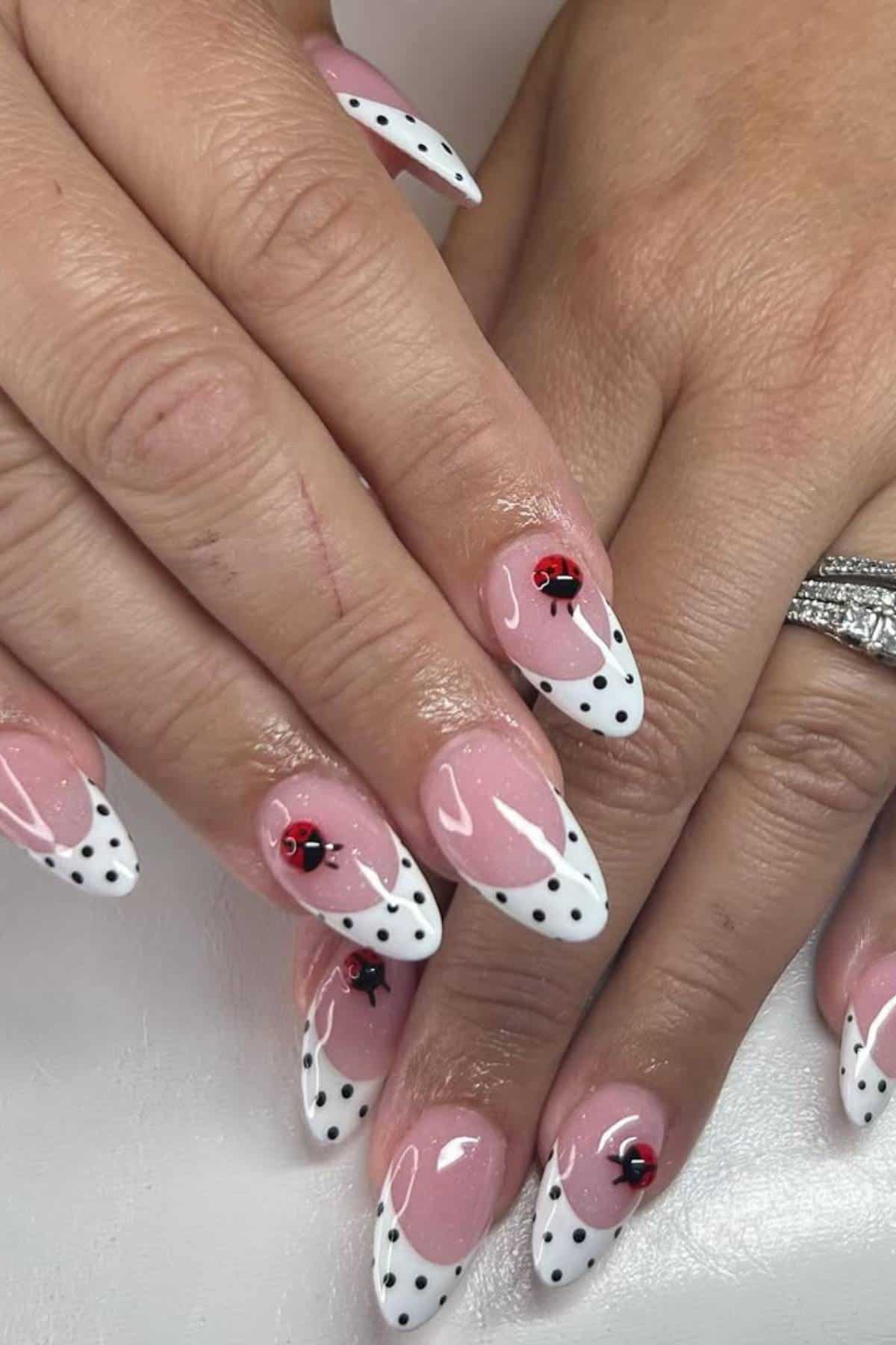 Hands with almond-shaped nails featuring white French tips, black polka dots, and small red ladybug designs; one hand has a diamond ring on the ring finger.