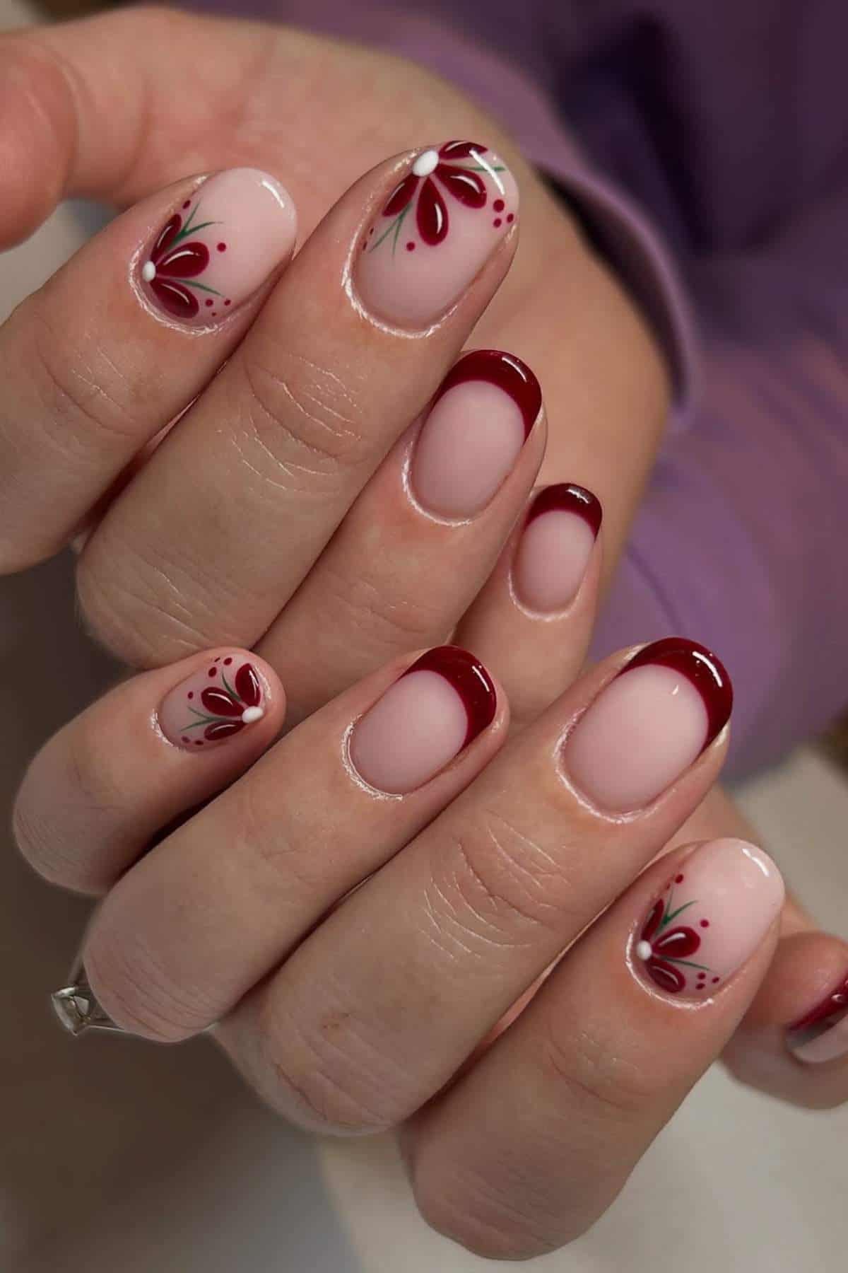 Hands with short oval nails featuring a light pink base, dark red French tips, and floral designs in red and green on select nails.