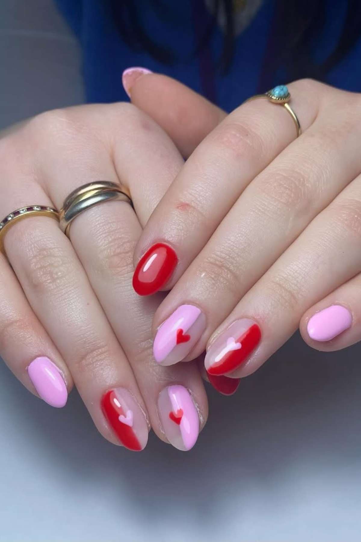 Hands with almond-shaped nails painted in pink and red polish, featuring small red heart designs on some nails. The person is wearing gold rings.