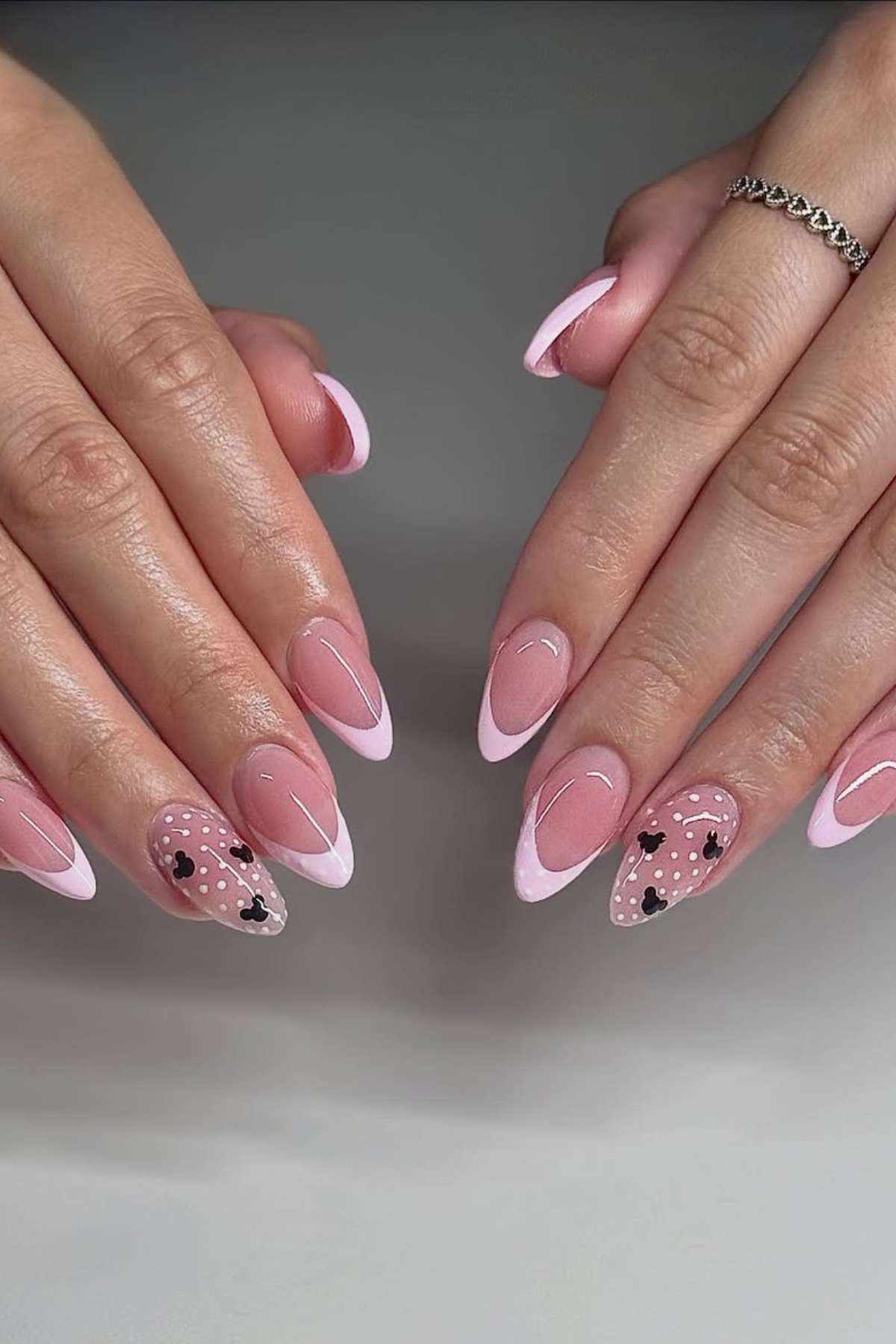 Hands with almond-shaped nails featuring a pink French tip design, with accent nails decorated with black hearts and white dots on a translucent pink base.