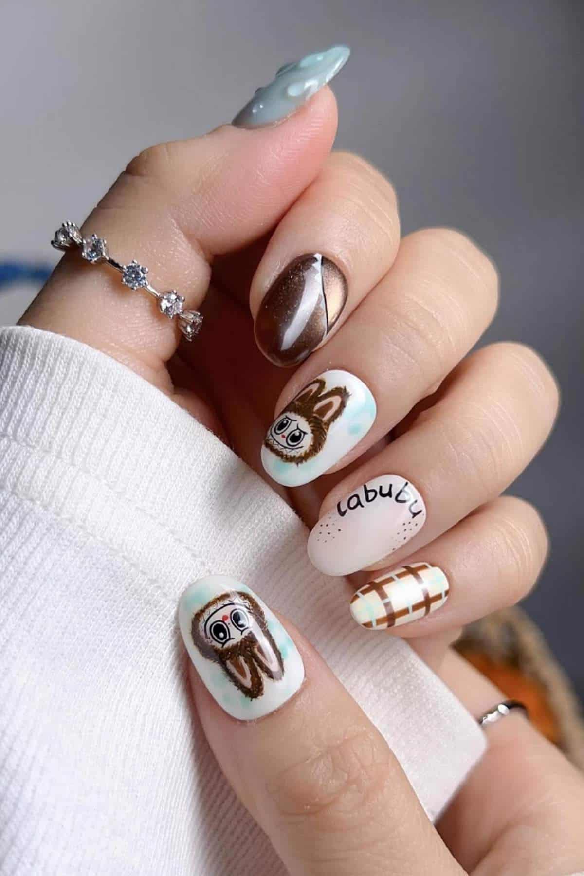 A hand with manicured nails features various designs, including a cartoon animal face, the word "cabubu," a brown swirl, plaid pattern, and light blue with 3D dots.