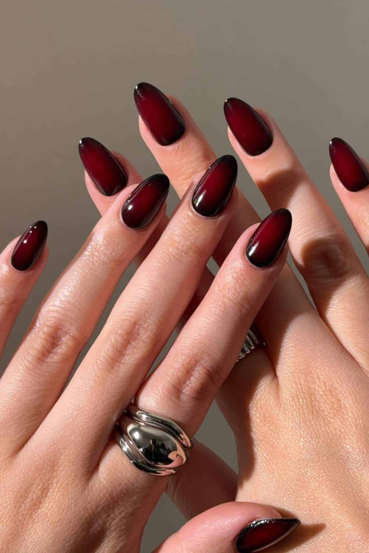 Close-up of hands with almond-shaped nails painted in a dark red ombre design; one hand wears a silver ring.