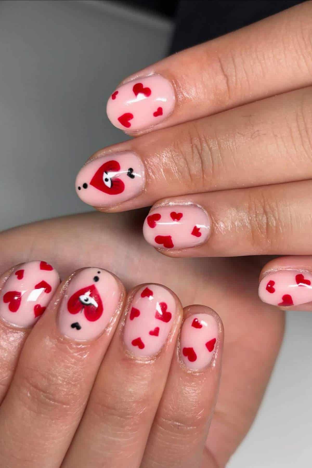 Fingernails painted with a pale pink base, decorated with small red hearts and two accent nails featuring larger heart designs with eyes and black dots.