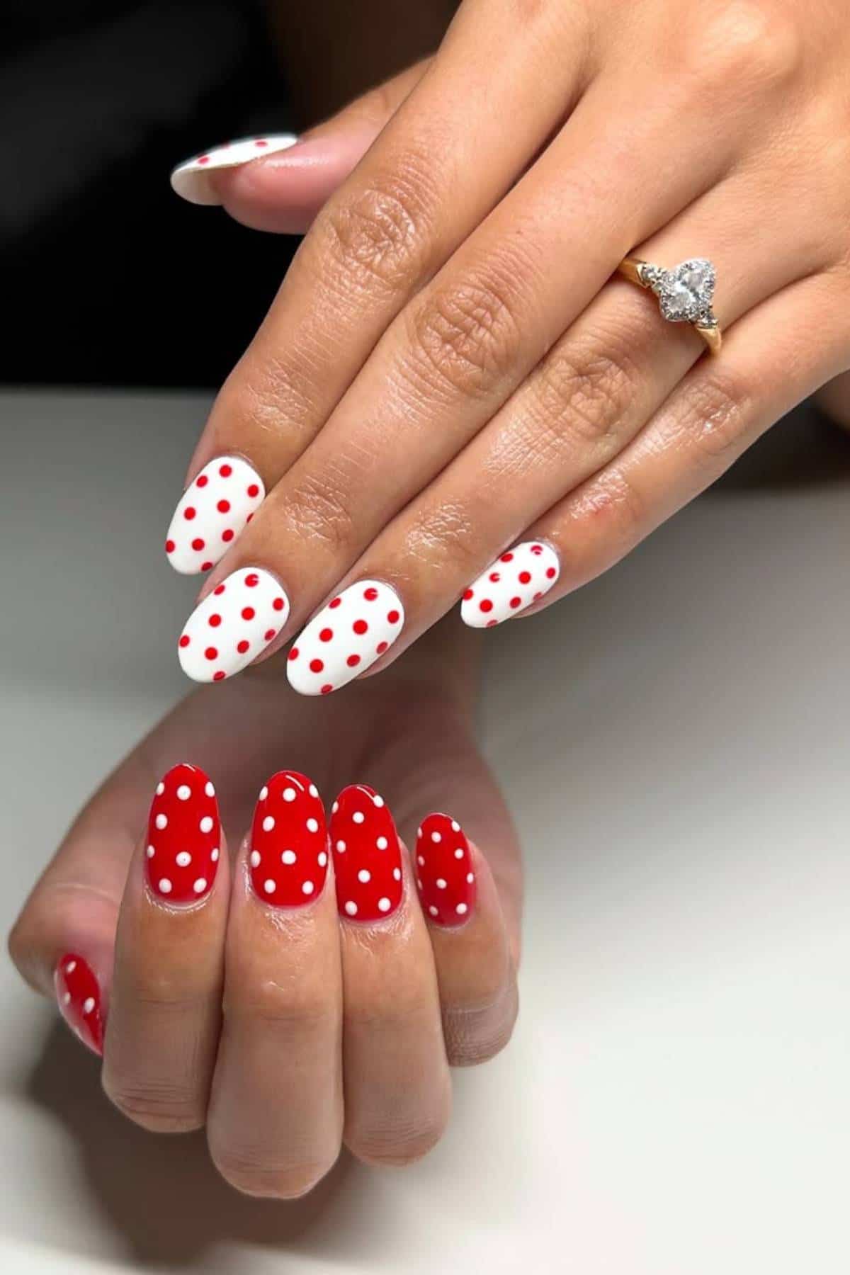 Hands with manicured nails; one hand has white nails with red polka dots, the other has red nails with white polka dots. A heart-shaped ring is on one finger.