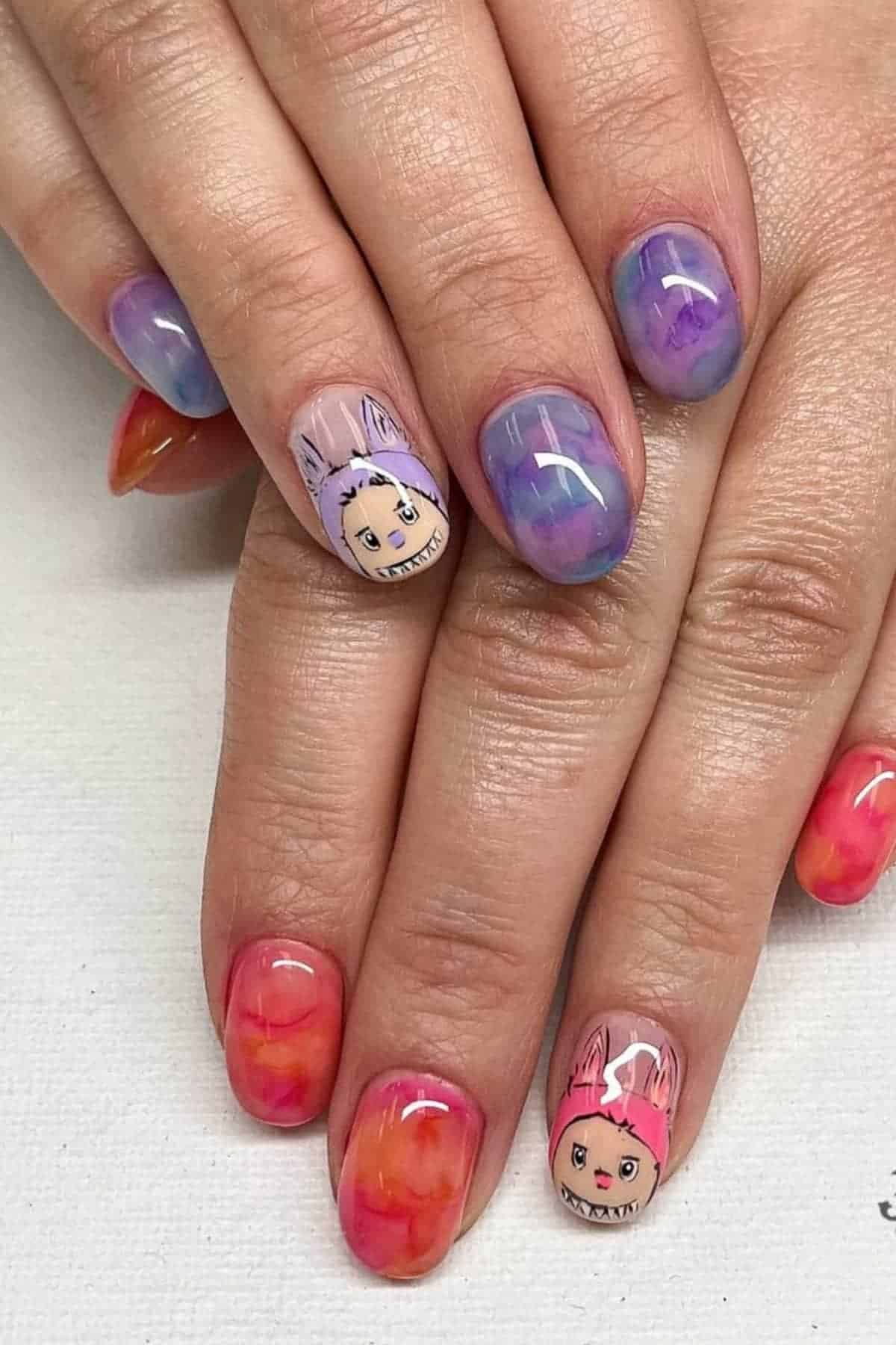 Hands with colorful gradient nail art; two nails feature cartoon faces with animal ear hoods, while others have purple, blue, orange, or pink abstract designs.