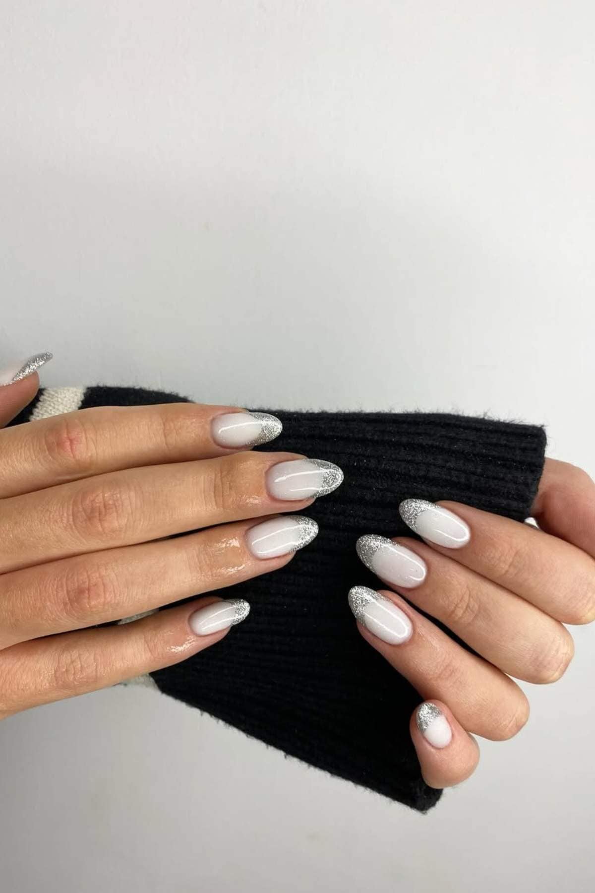 A hand with almond-shaped nails featuring a sheer pink base and silver glitter tips, resting on a black ribbed sleeve against a white background.
