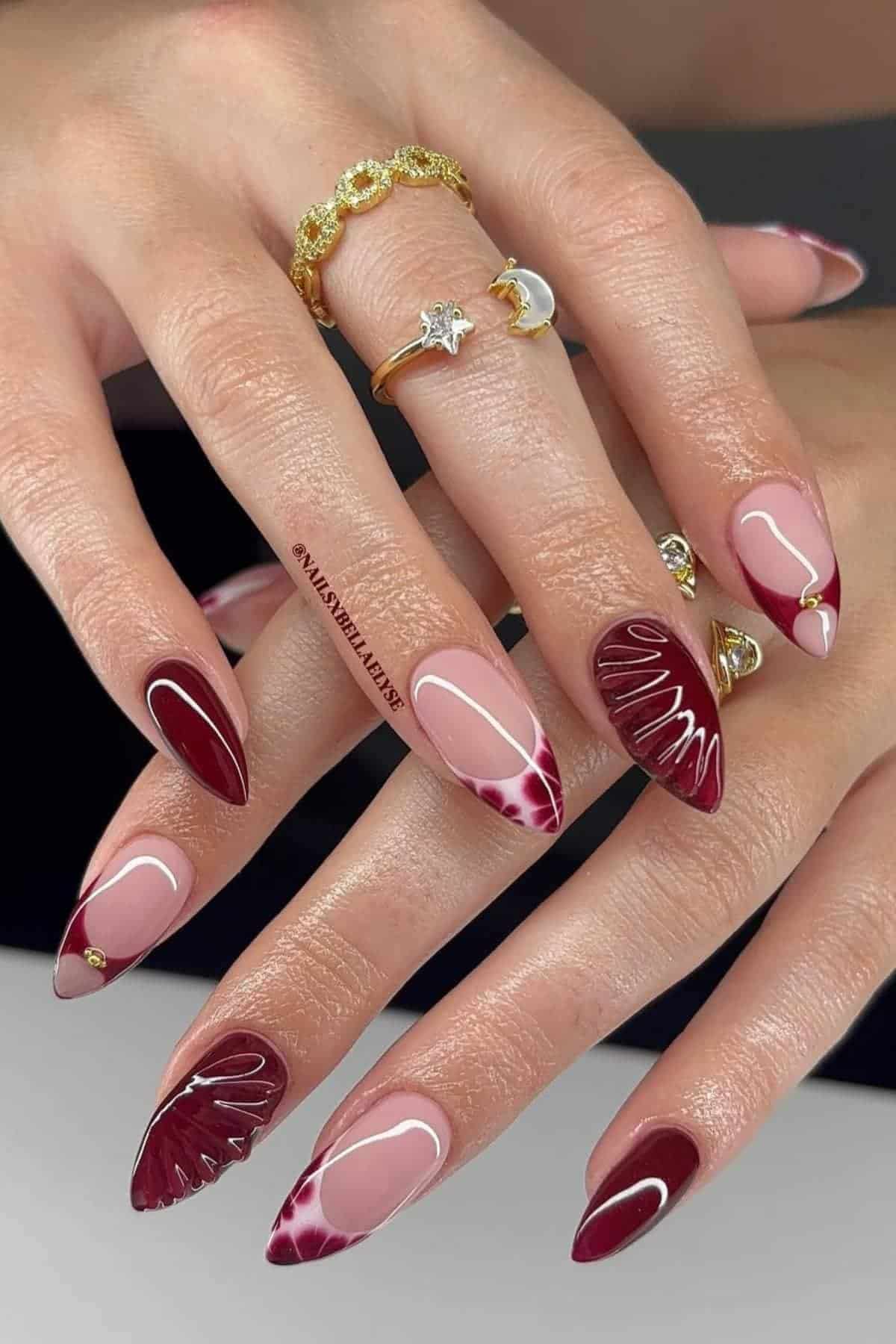 Close-up of hands with almond-shaped nails featuring burgundy, white, and nude abstract nail art designs; wearer has gold rings on two fingers.