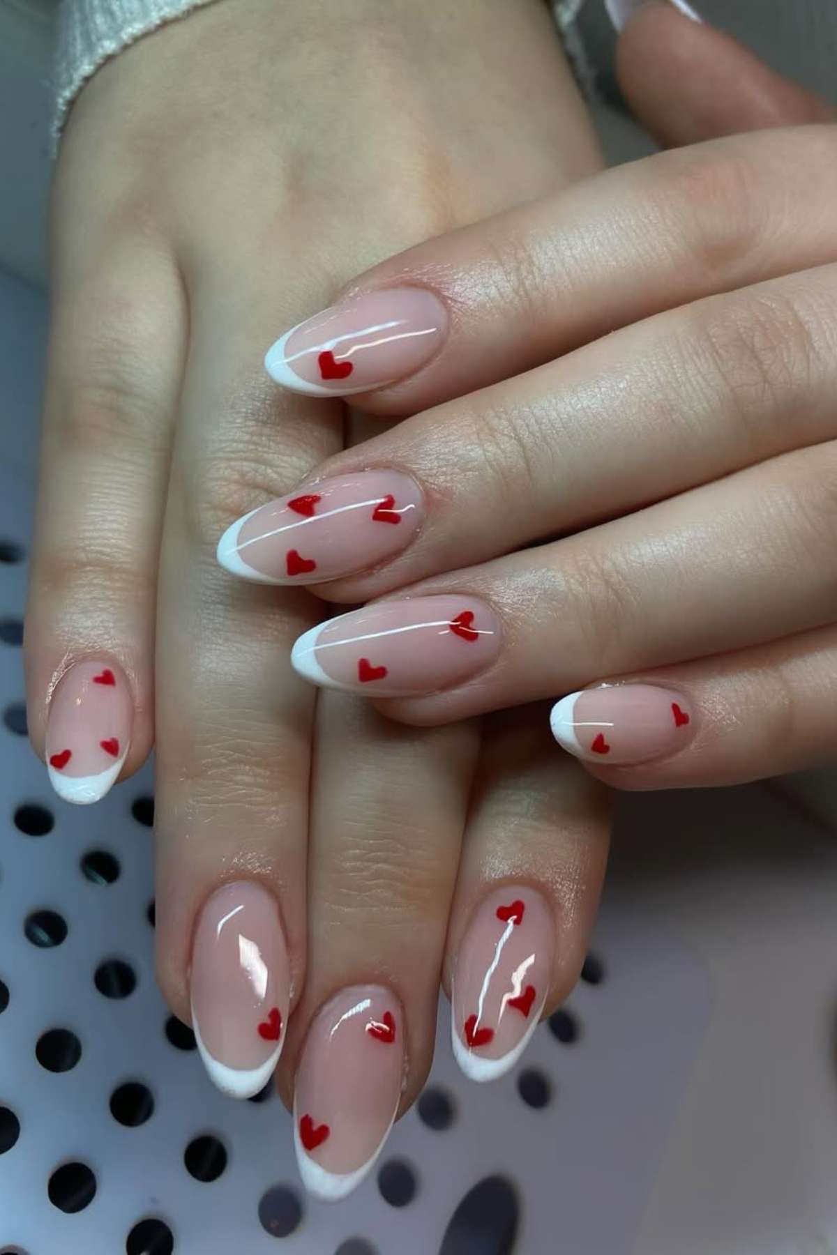 Hands with almond-shaped nails featuring French tips and small red heart decorations on a nude base.