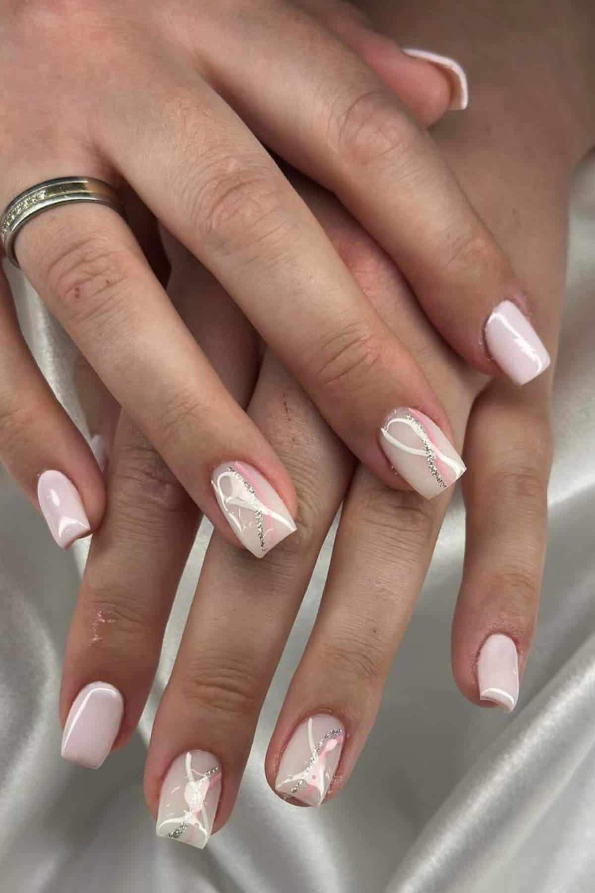 Close-up of hands with short, pale pink glossy nails featuring delicate silver and white ribbon-like designs on two accent nails.
