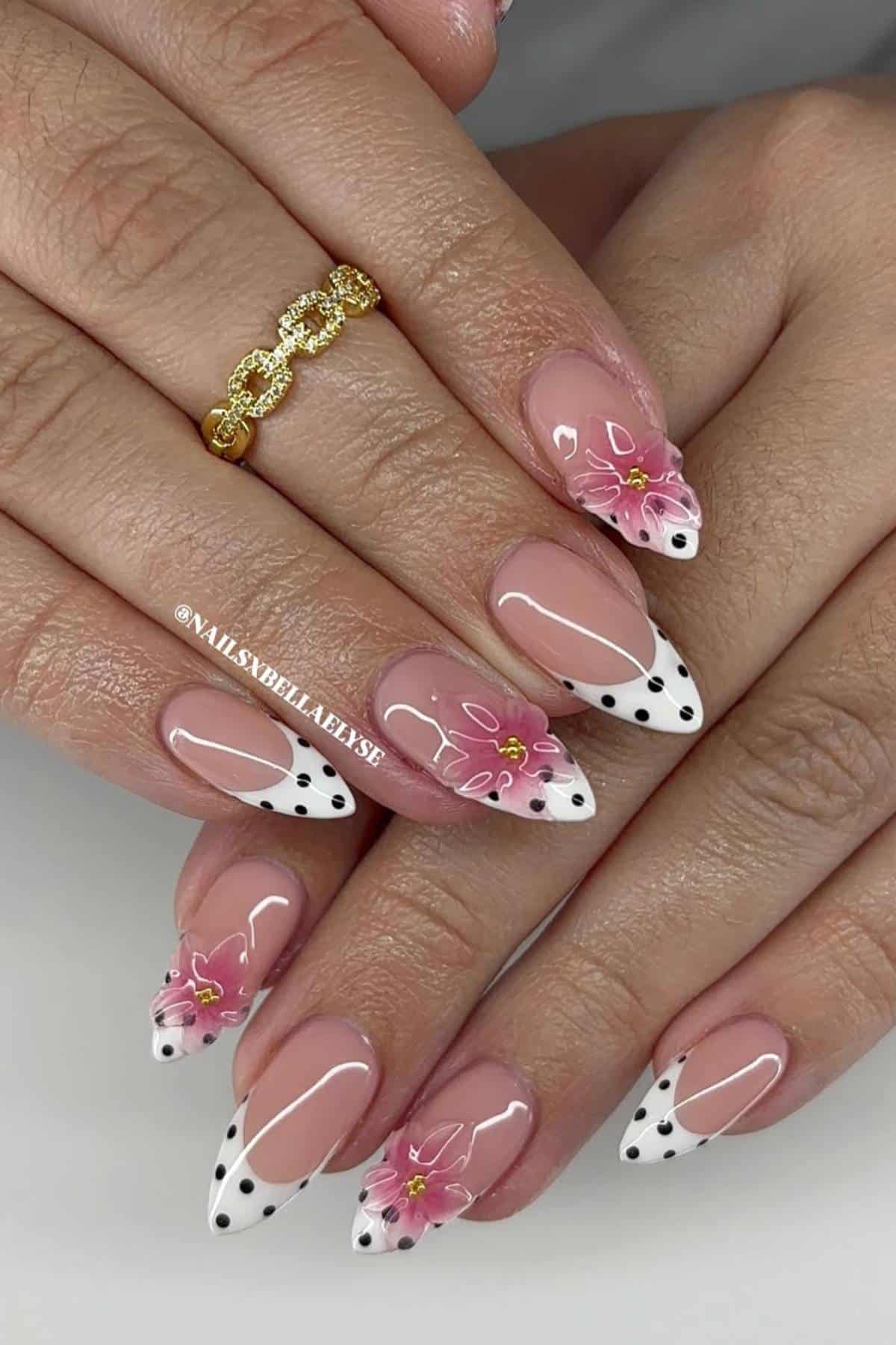 Hands with almond-shaped nails featuring white French tips with black polka dots and pink 3D floral designs, shown with a gold ring on one finger.