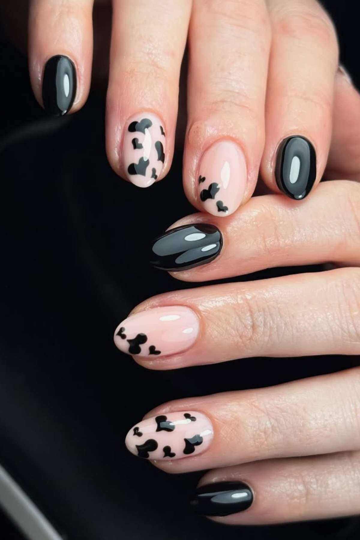 Hands with oval nails featuring alternating black polish and nude polish with black abstract cow print designs.