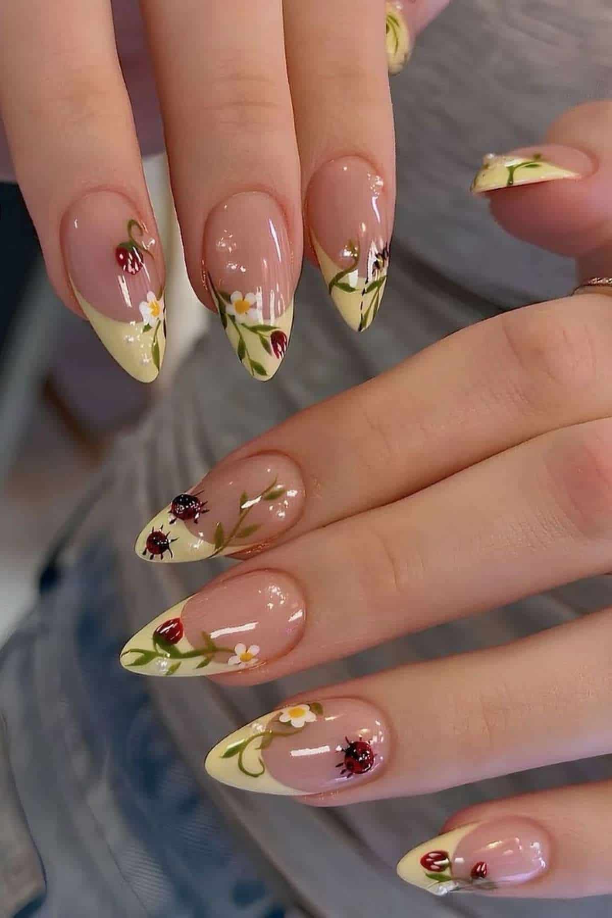 Close-up of hands with almond-shaped nails featuring pale yellow French tips, floral designs, and small ladybug accents on a natural pink base.