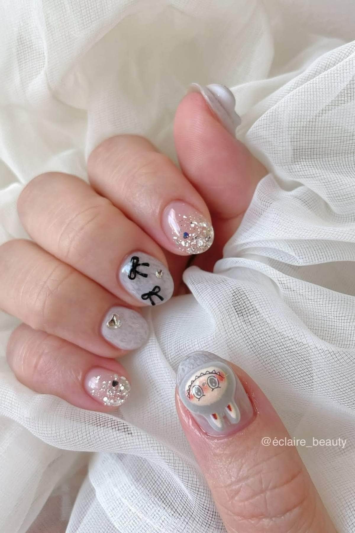 A hand with short, almond-shaped nails featuring silver glitter, small gems, black letters "R," and a cartoon face design on the thumb, resting on white fabric.
