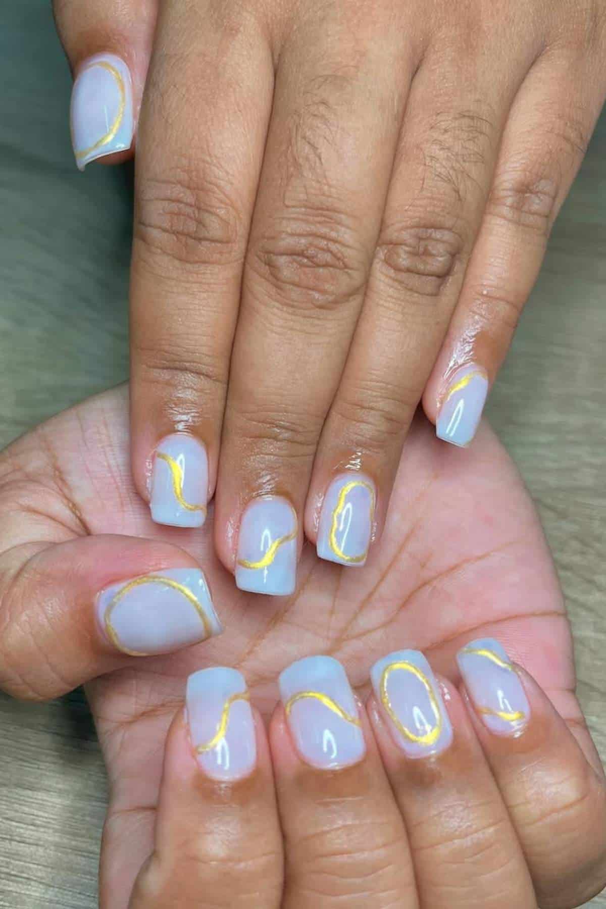 Hands with short, square-shaped nails painted light blue and decorated with a thin, wavy gold line on each nail.