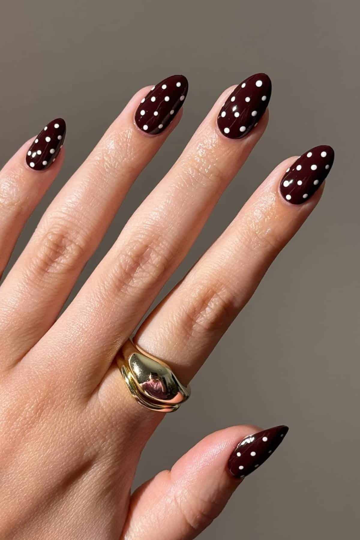 A hand with almond-shaped nails painted dark brown with white polka dots, wearing two gold rings on the middle finger, against a neutral background.