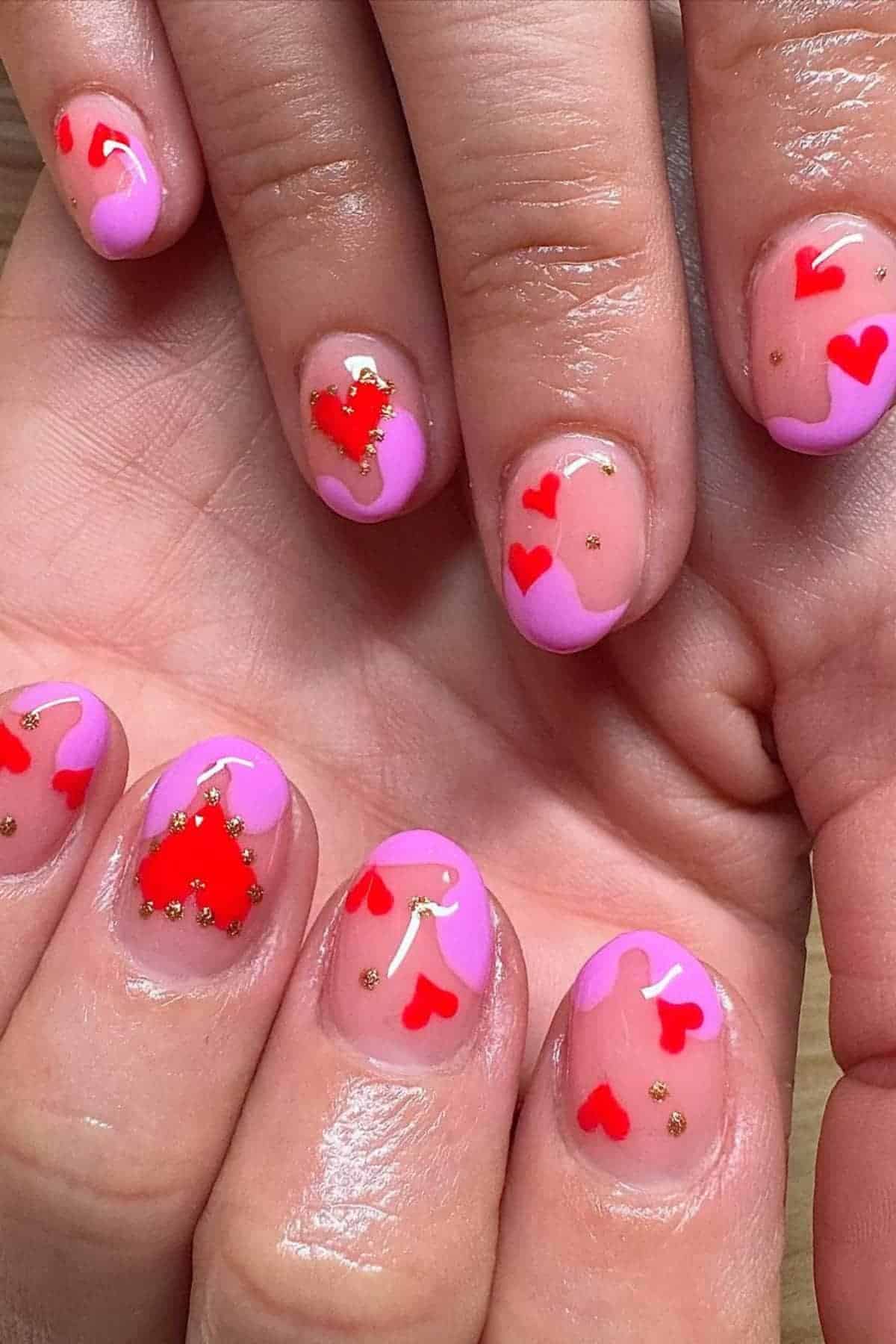 Close-up of hands with manicured nails featuring pink tips, red and pink heart designs, and small gold accents on a nude base.